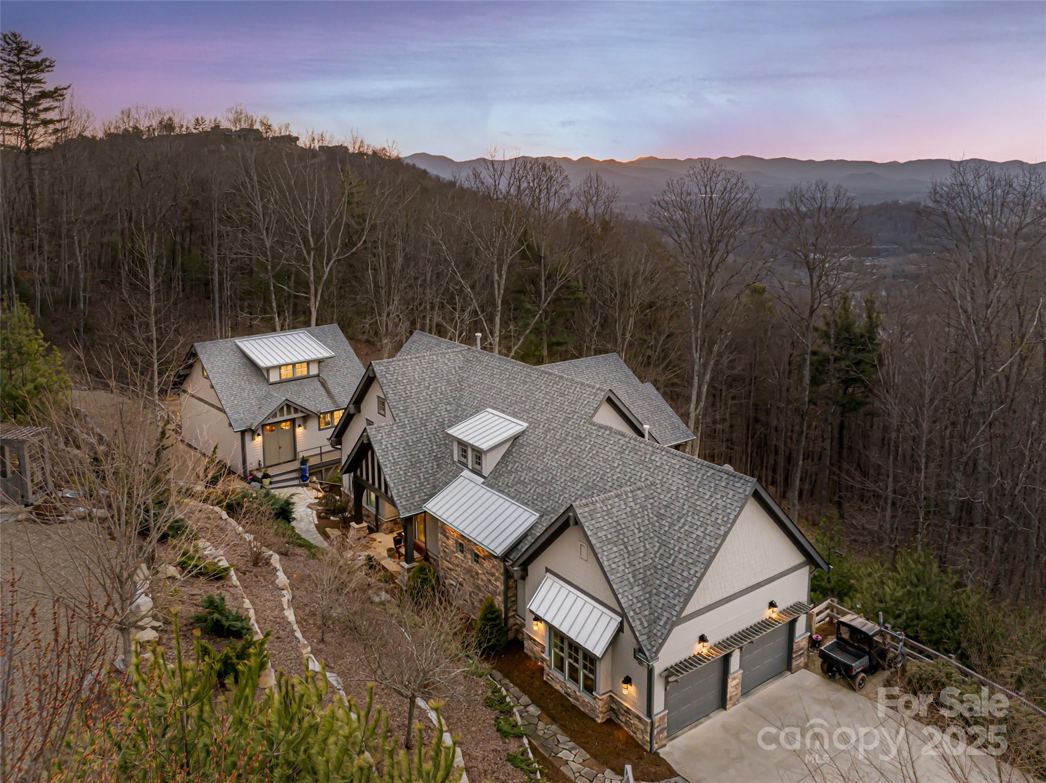 Hillside home with illuminated windows, surrounded by leafless woods and overlooking distant mountain ridges.
