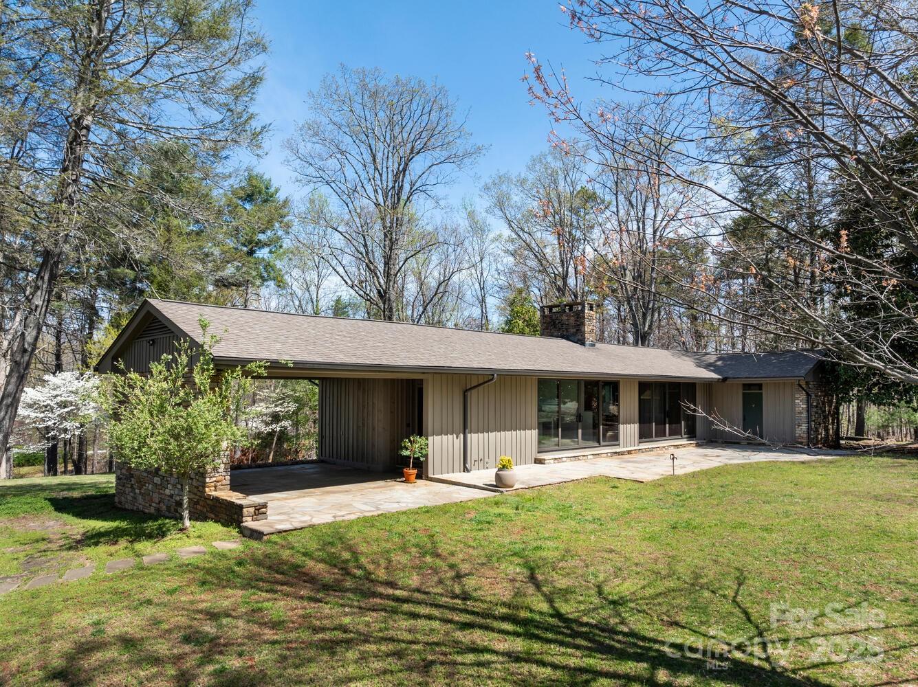 Single-story home with a stone patio and low stone planter wall, surrounded by mature trees.