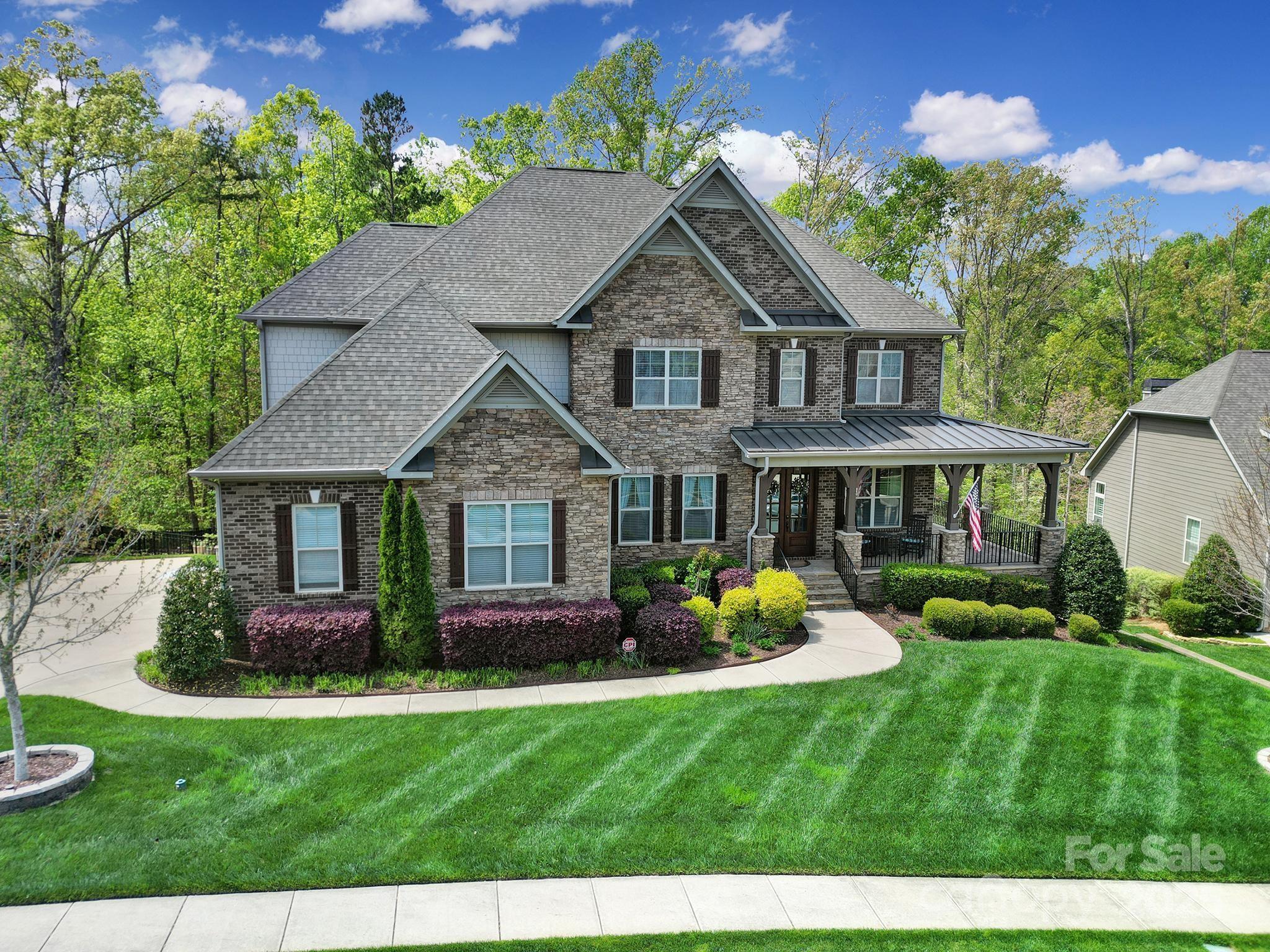 Two-story stone-and-brick house with a covered porch, landscaped shrubs, and a broad green lawn.