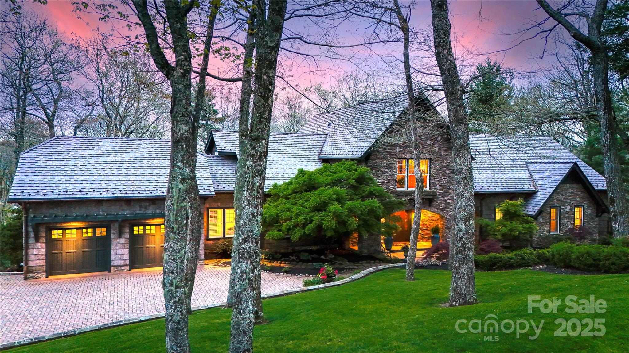 Stone house with a slate roof, glowing windows, and brick driveway beneath tall trees at dusk.
