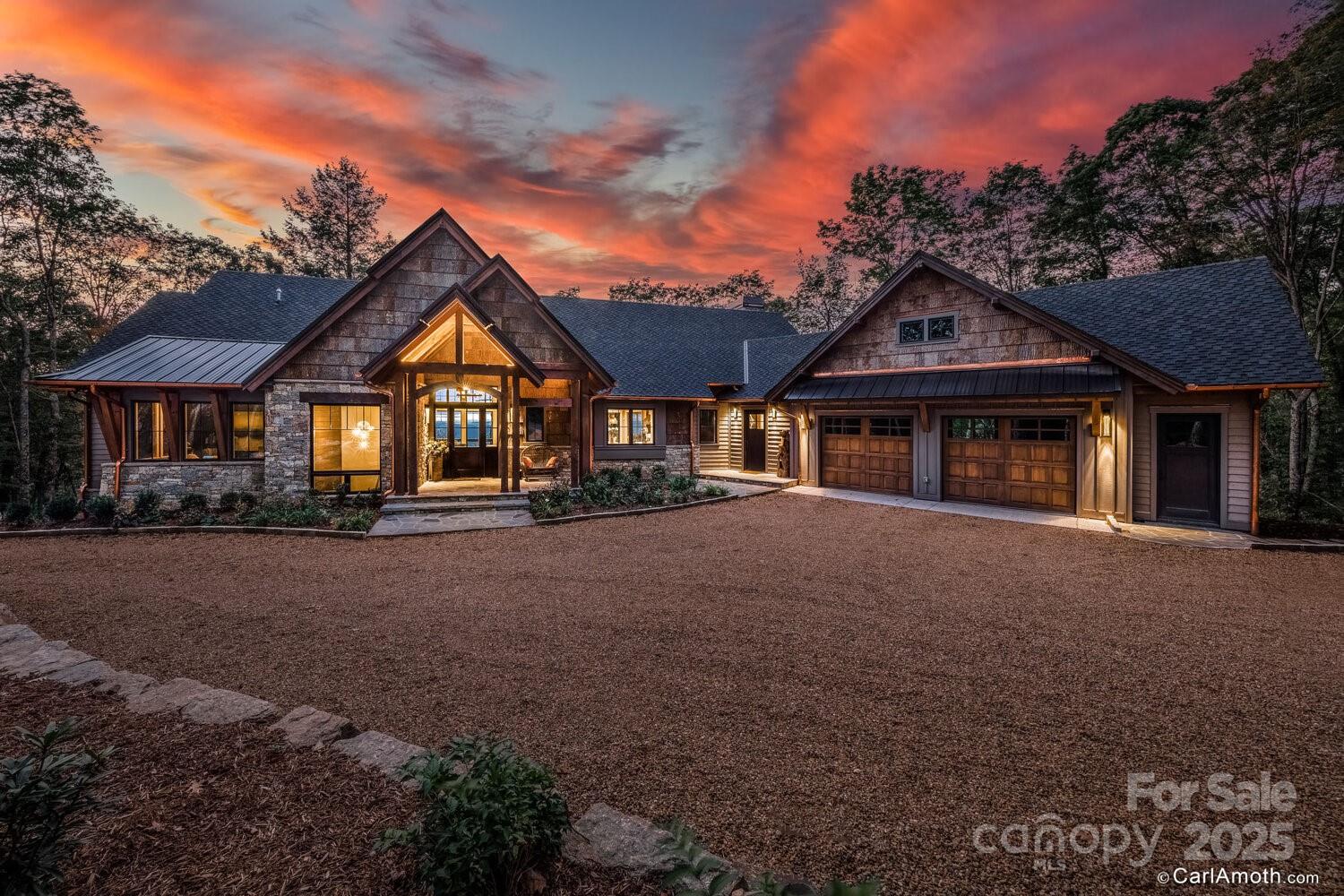 Wood-and-stone home with a broad gravel drive and twin garage doors beneath a vivid sunset sky.