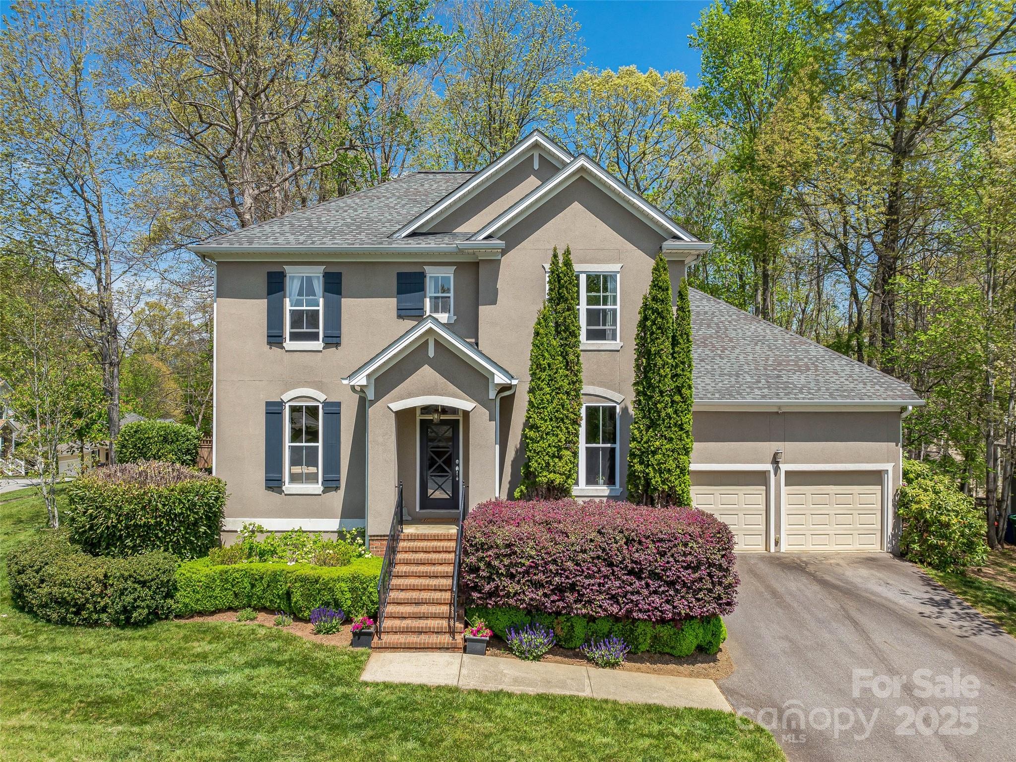 Two-story beige home with blue shutters, brick entry steps, landscaped shrubs, and a two-car garage.