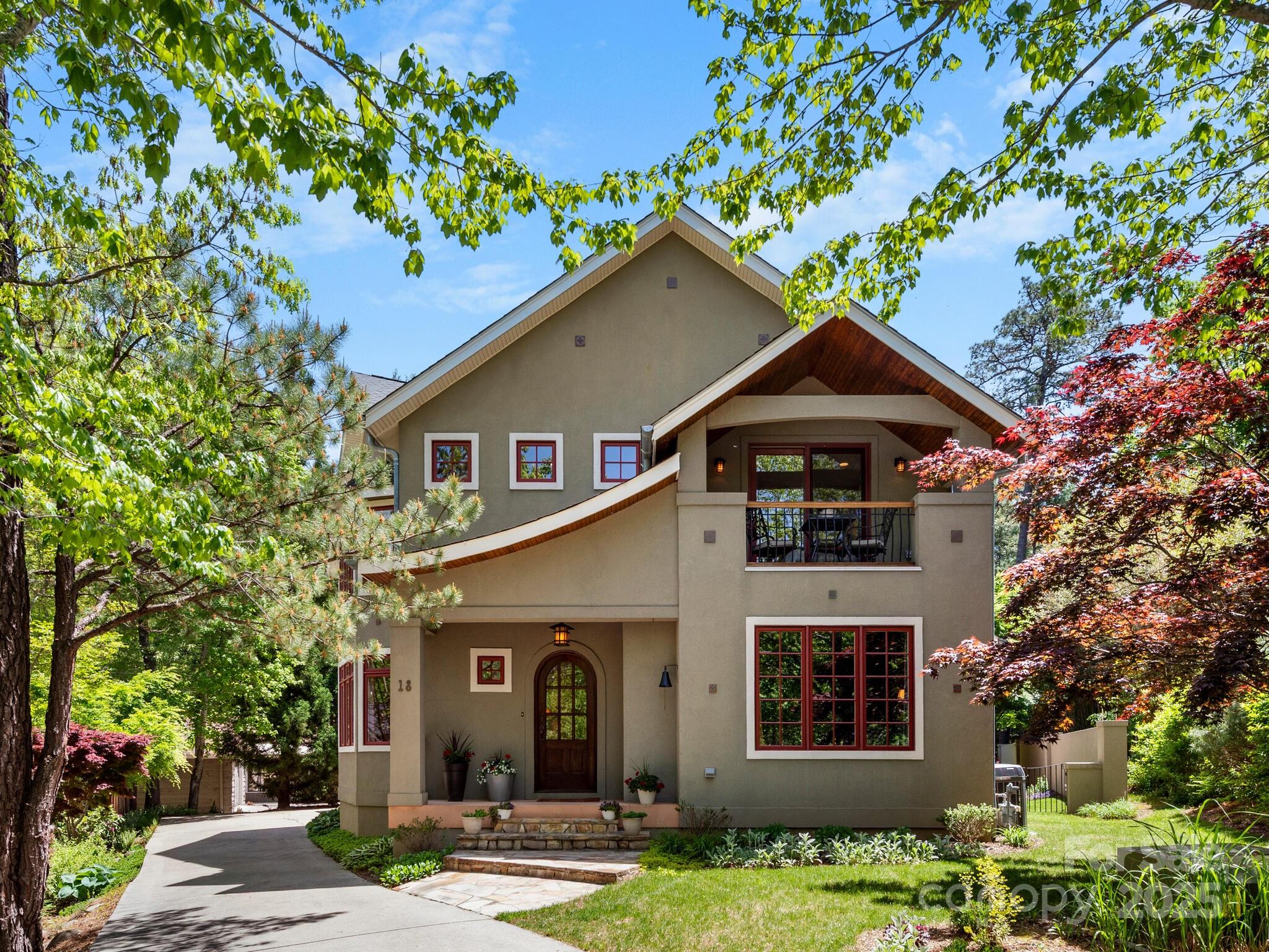 Two-story beige home with red-trimmed windows, a covered balcony, and mature trees framing the front yard.