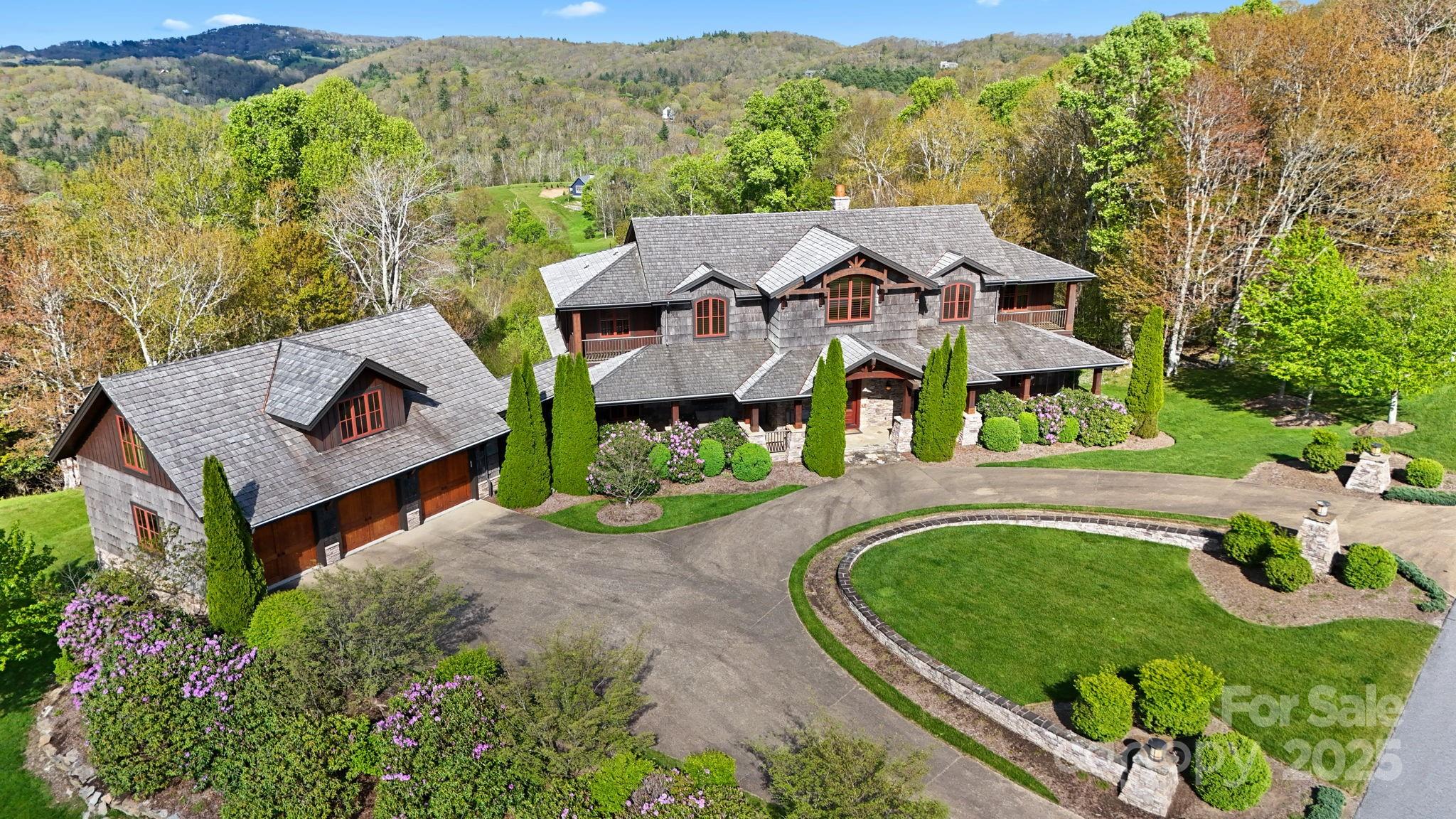 Aerial view of a large shingle-sided home with a circular driveway, landscaped gardens, and wooded mountain surroundings.