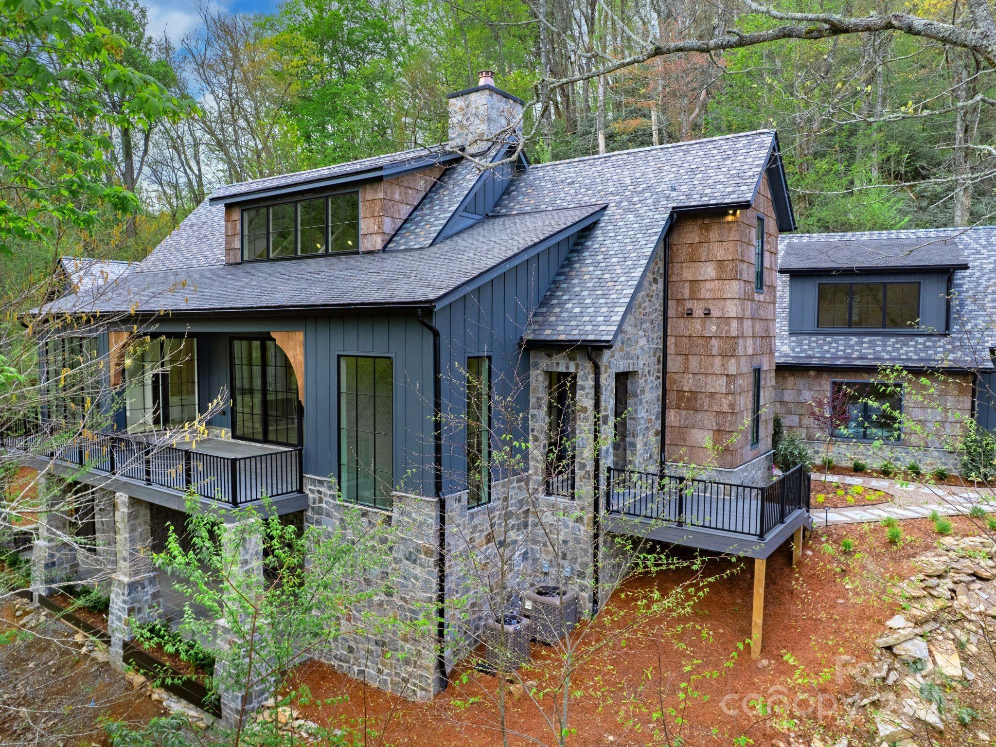 Modern stone-and-cedar home nestled among trees, with steep gray roofs and raised decks.