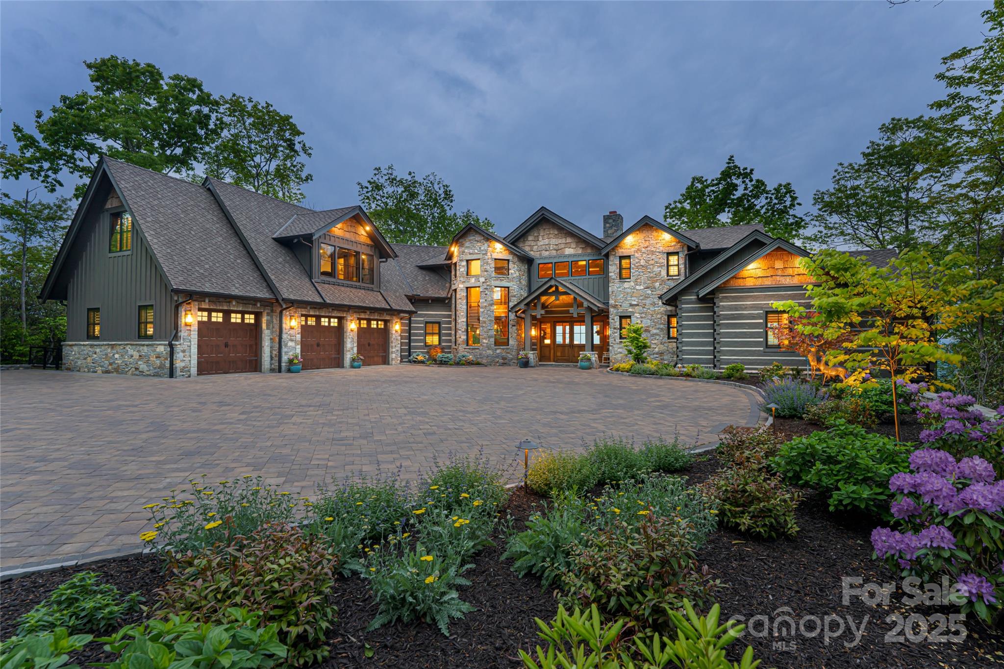 Stone-and-siding home with a broad paved driveway, warmly lit windows, and landscaped flower beds at dusk.