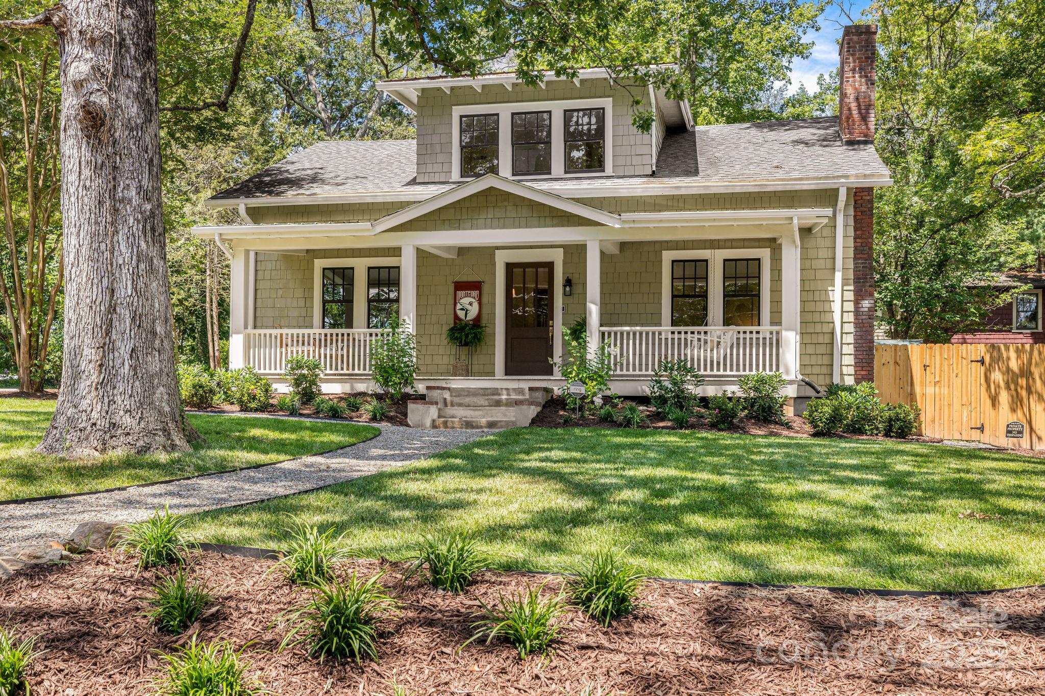 Gray-shingled two-story house with a white-railed porch, brick chimney, and landscaped yard beneath a large tree.