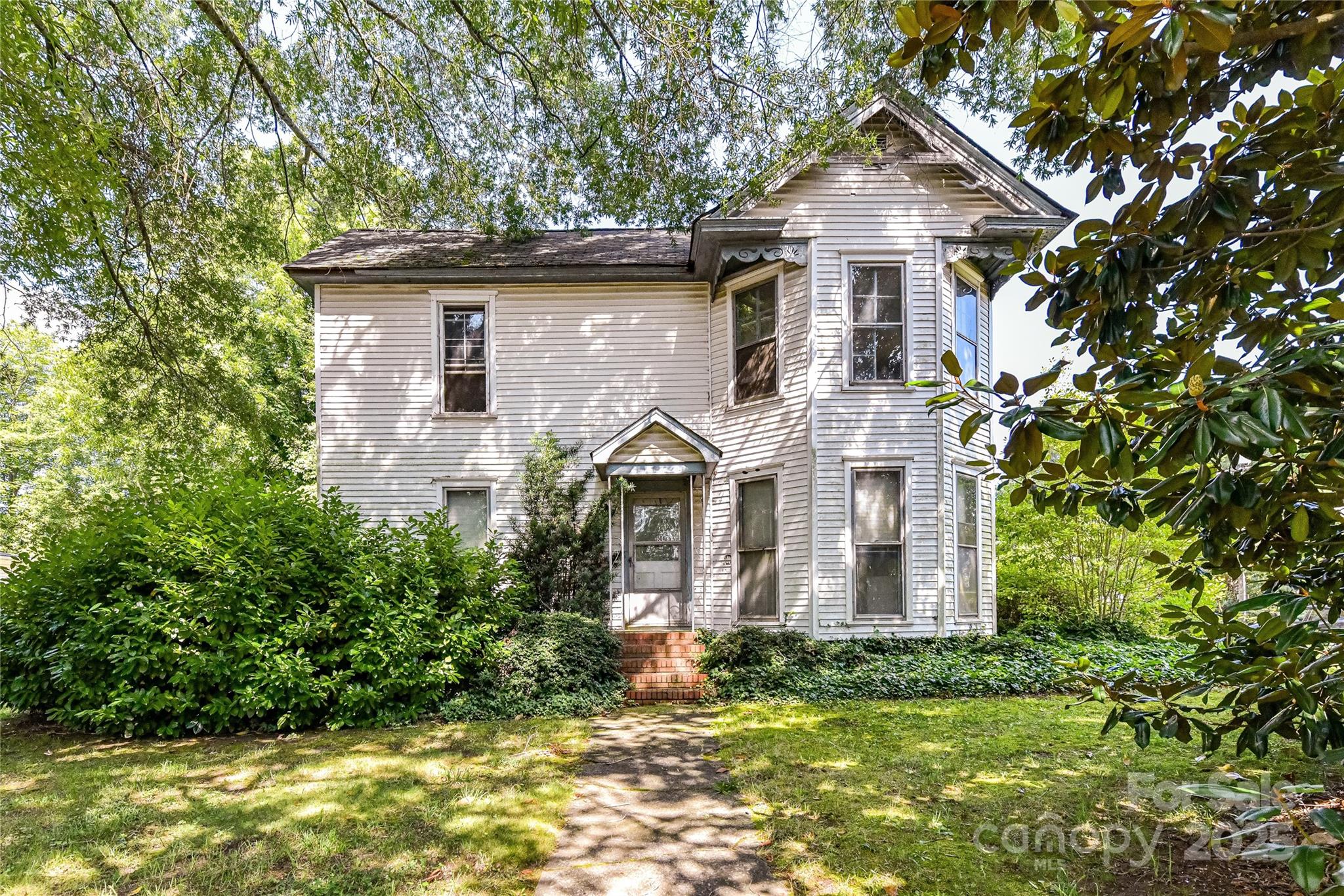 White two-story house with a projecting bay, brick steps, and a leafy front yard.