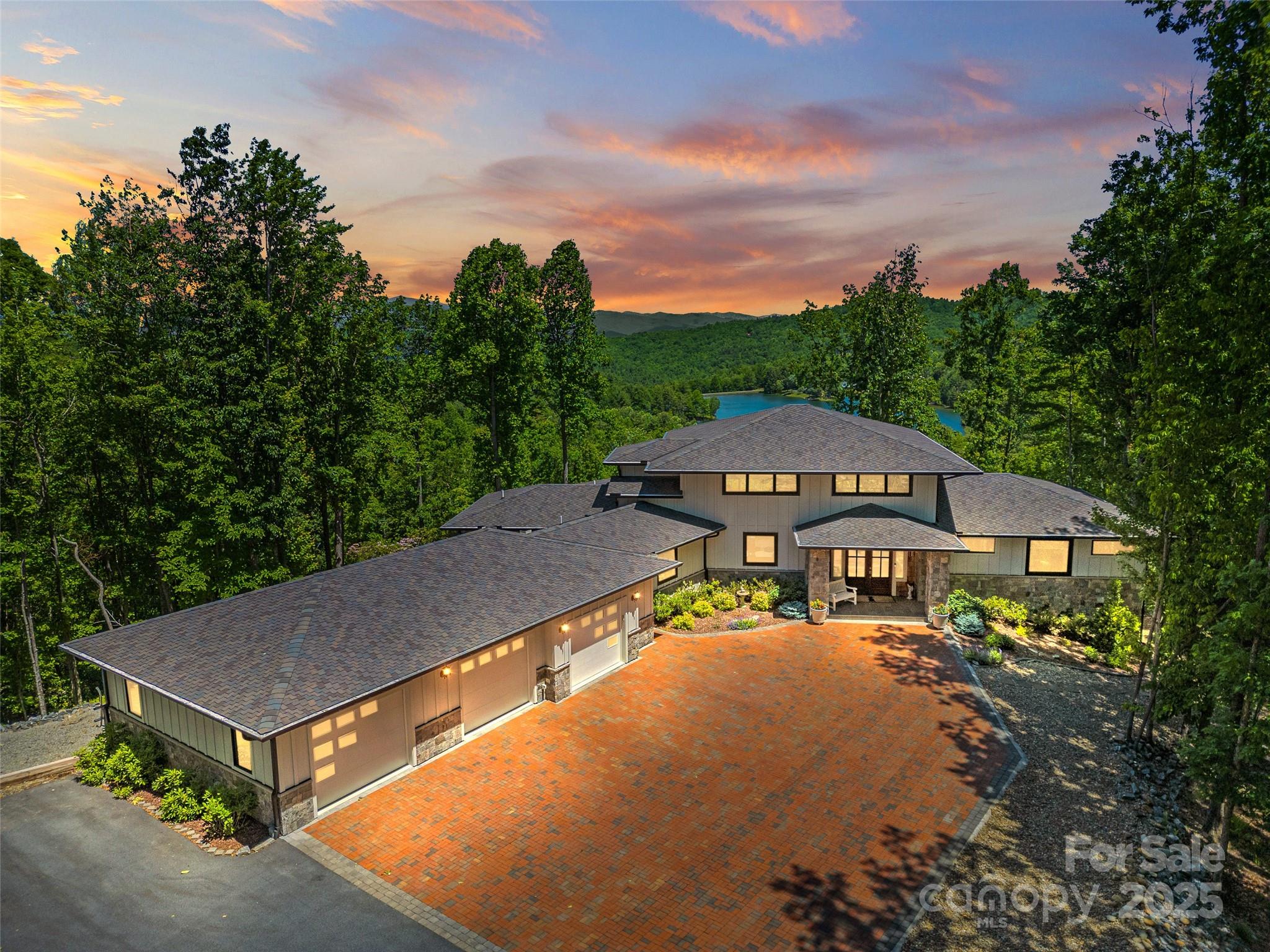 Modern home with a broad brick driveway, surrounded by forest, with a lake and mountains beyond.