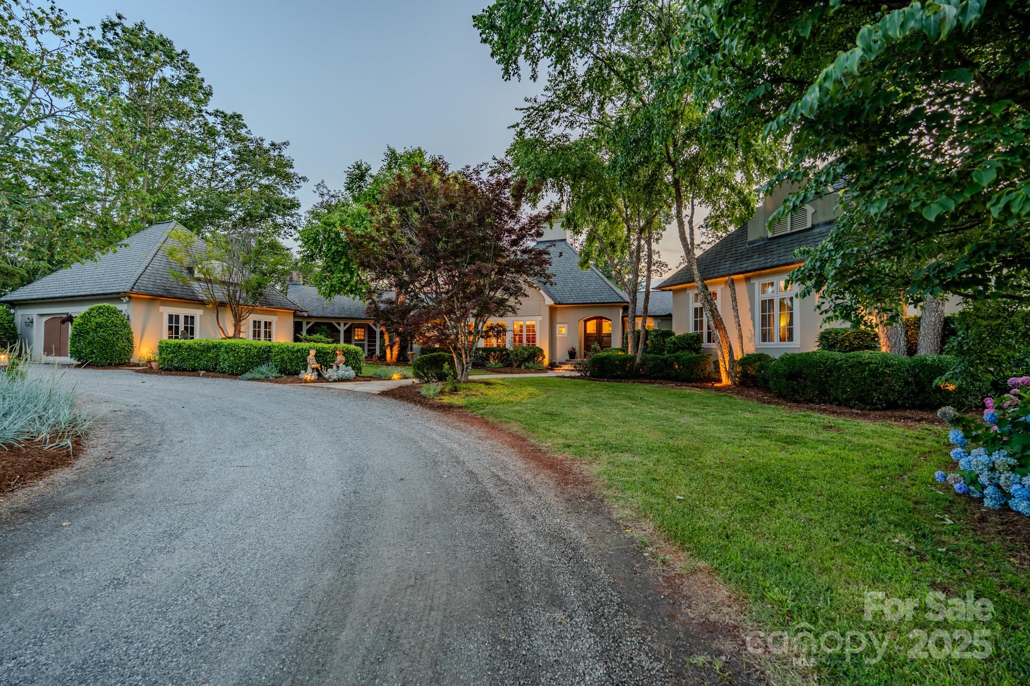 Large cream-colored home framed by mature trees, with a curved gravel driveway and warmly lit windows.