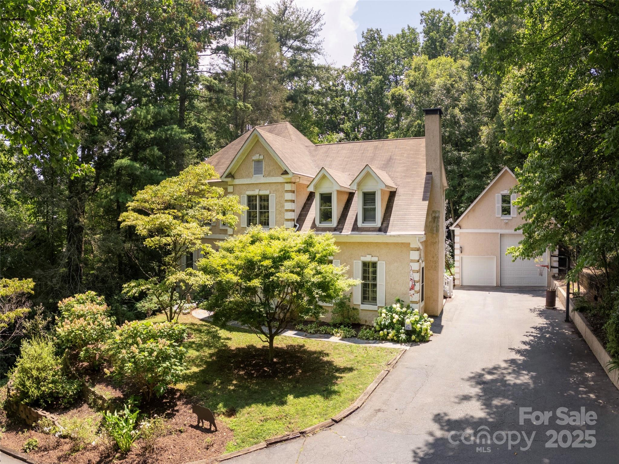 Beige two-story home with dormer windows, landscaped gardens, and a long driveway beneath mature trees.