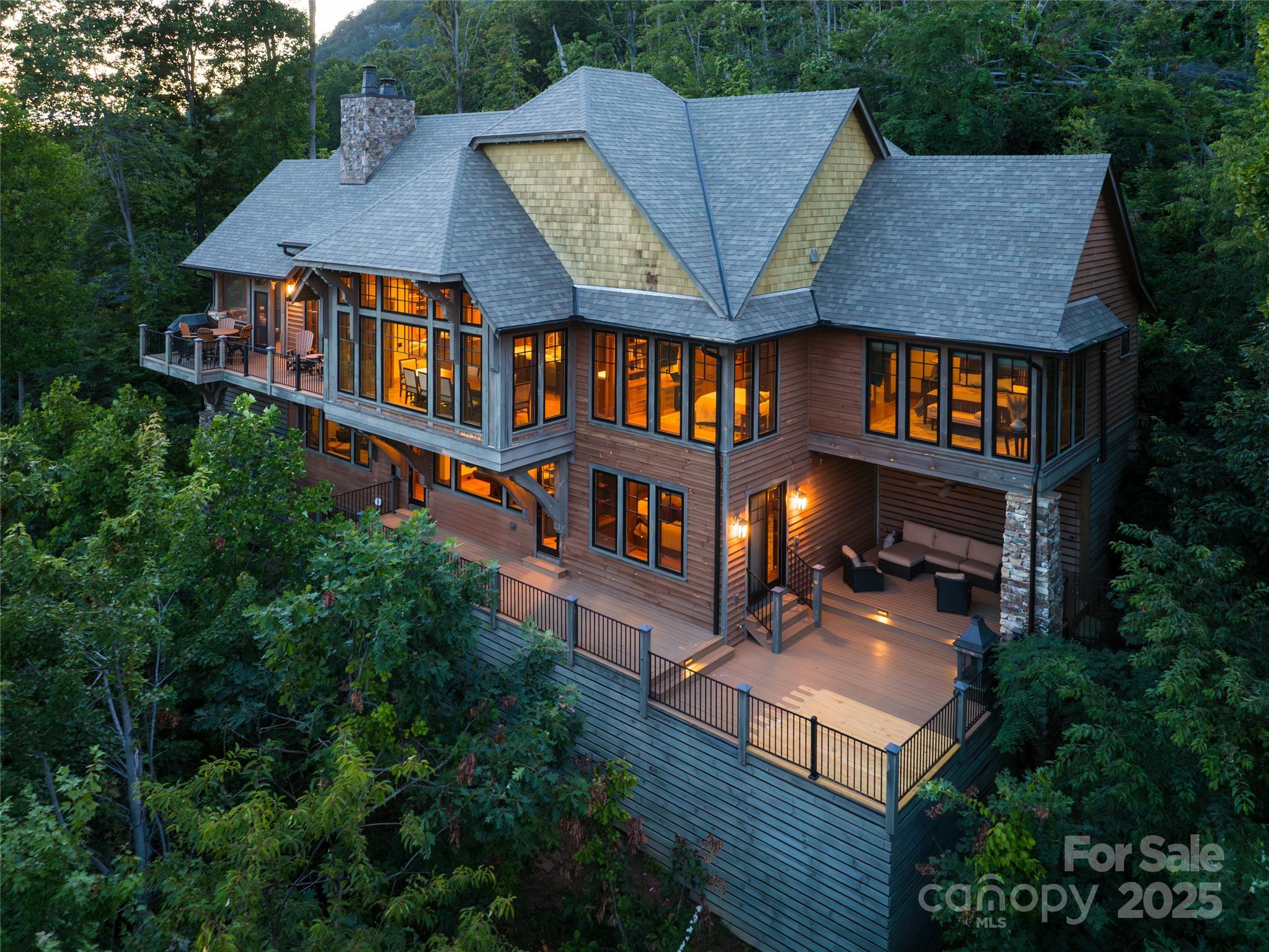 Wood-sided forest home with glowing windows and expansive decks, viewed from above at dusk.