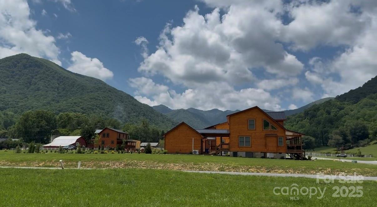 Wooden mountain homes across a grassy field beneath forested ridges and a partly cloudy sky.
