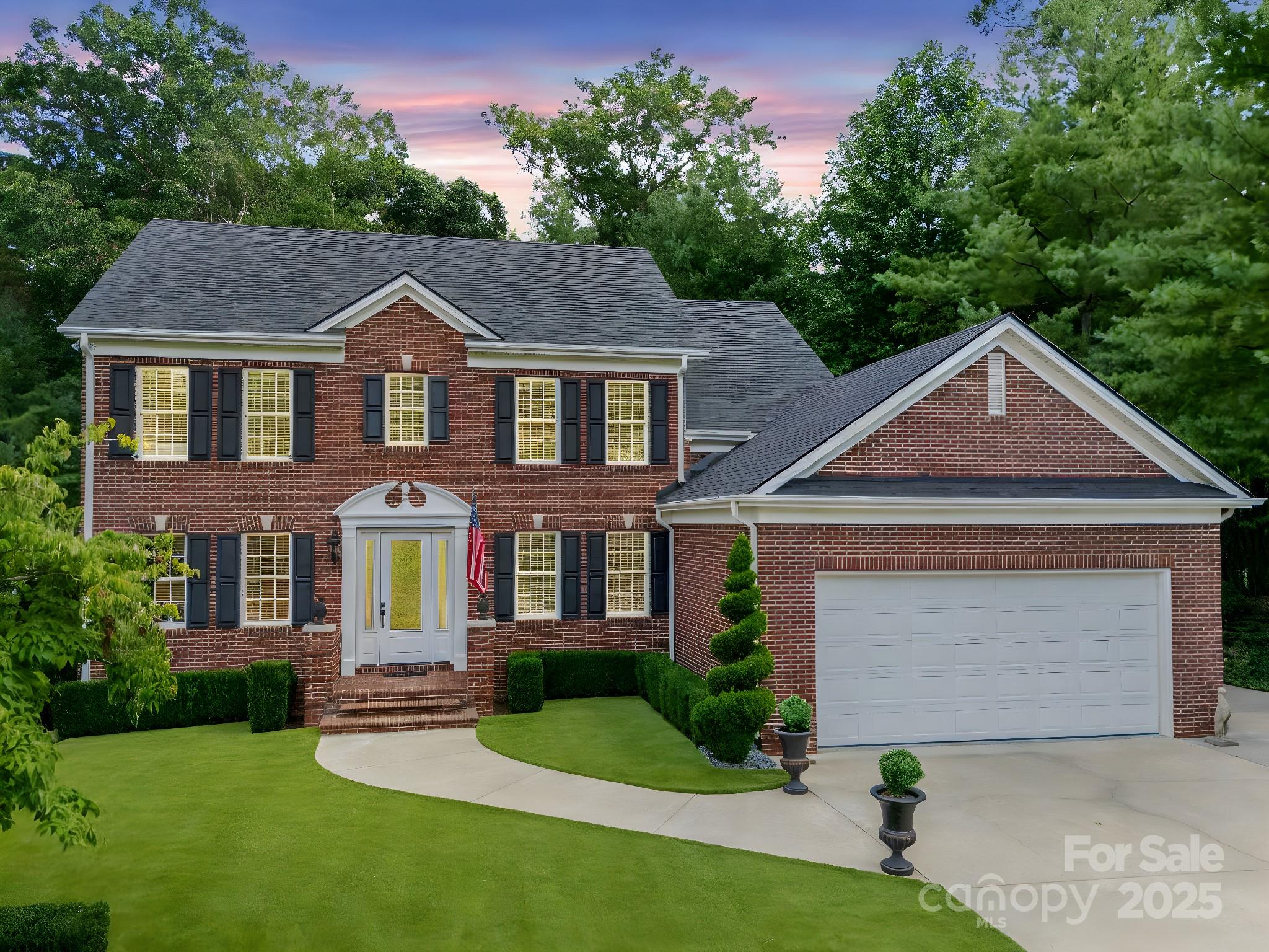 Two-story red brick house with black shutters, a two-car garage, and a landscaped lawn at dusk.