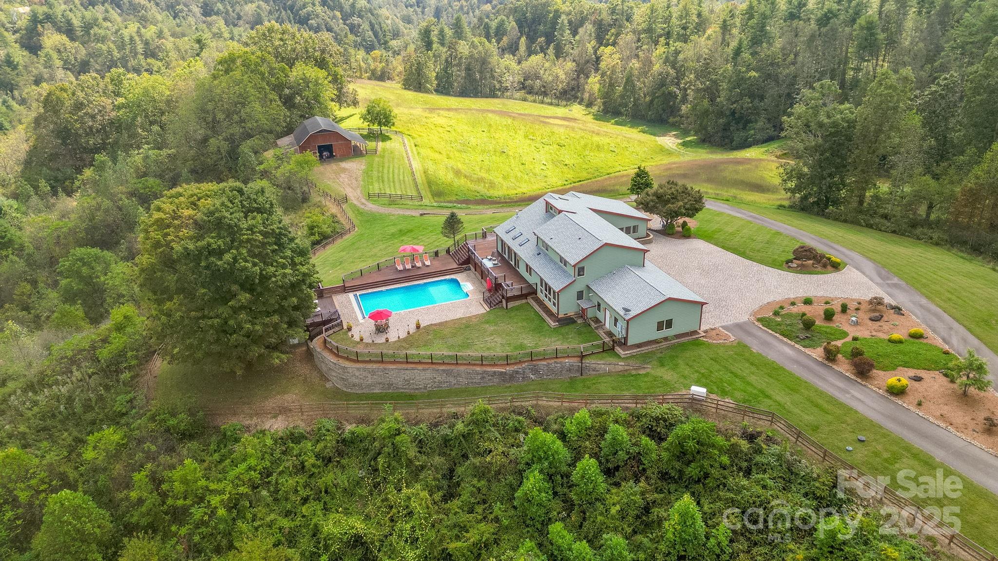 Aerial view of a country home with a pool, barn, fenced pasture, and wooded hills.