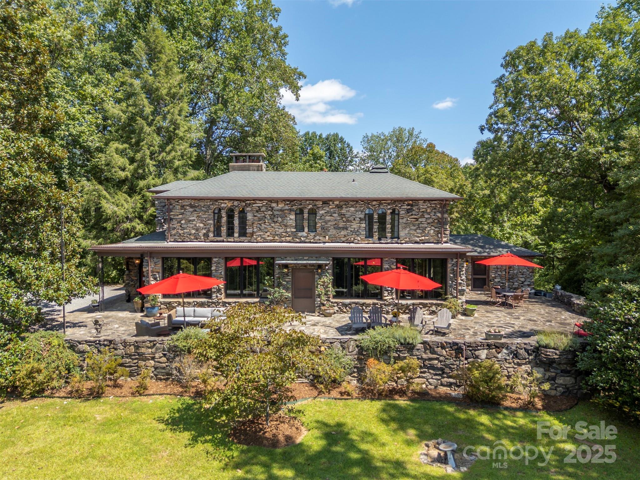 Two-story stone house with a spacious patio, red umbrellas, and mature trees.