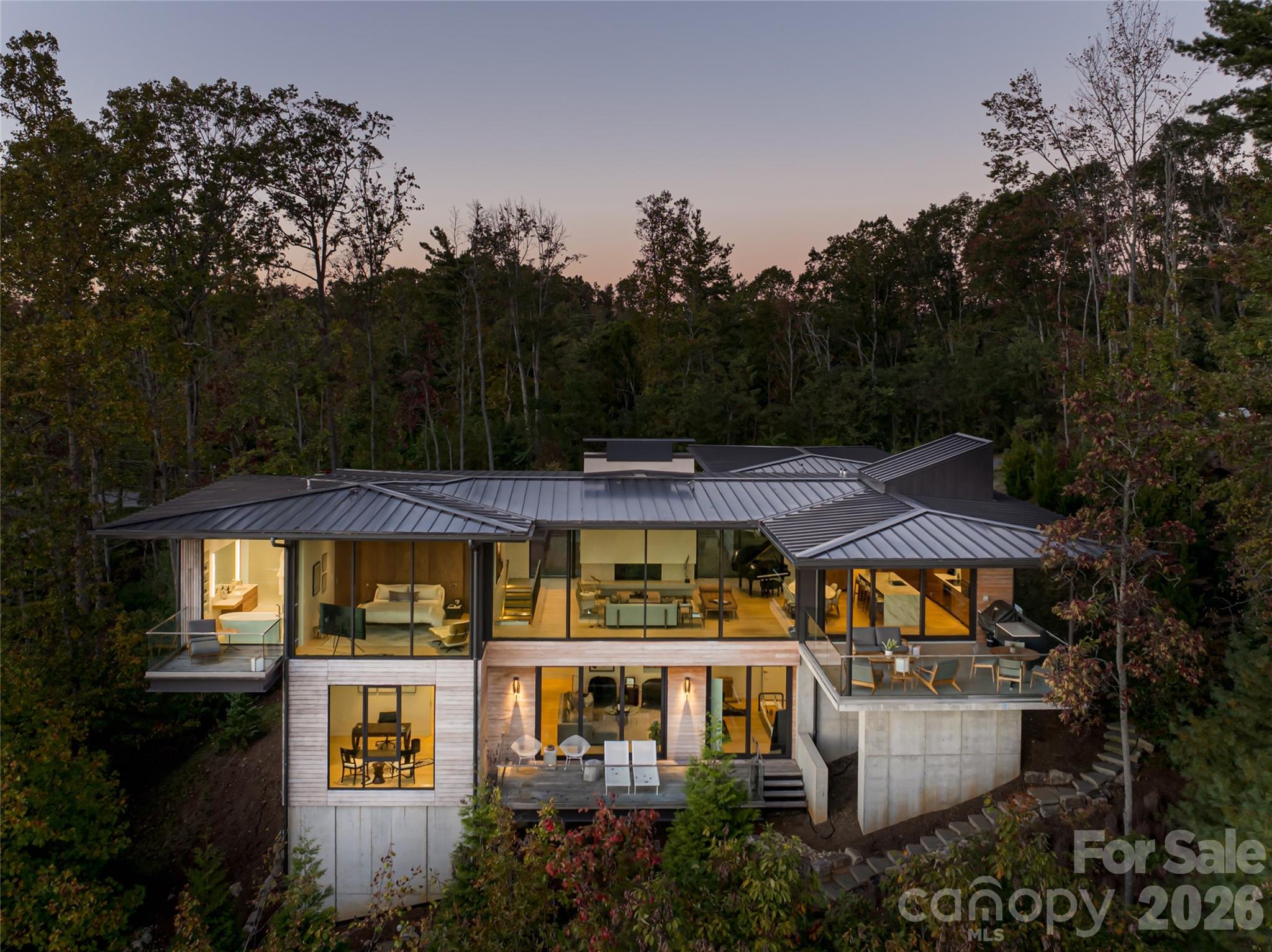 Modern glass-and-wood home with a metal roof and illuminated interiors, surrounded by trees at dusk.