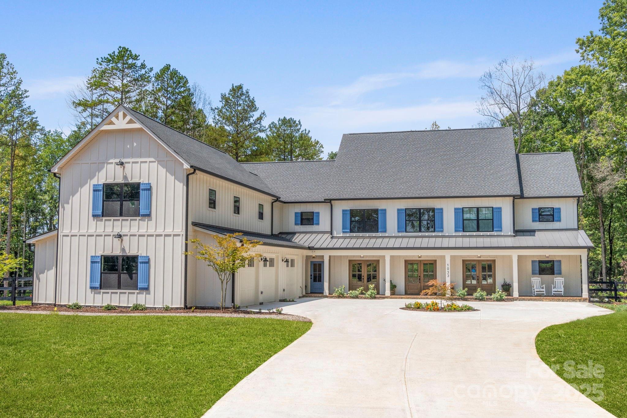 Large light-colored farmhouse with blue shutters, a broad curved driveway, and mature trees.