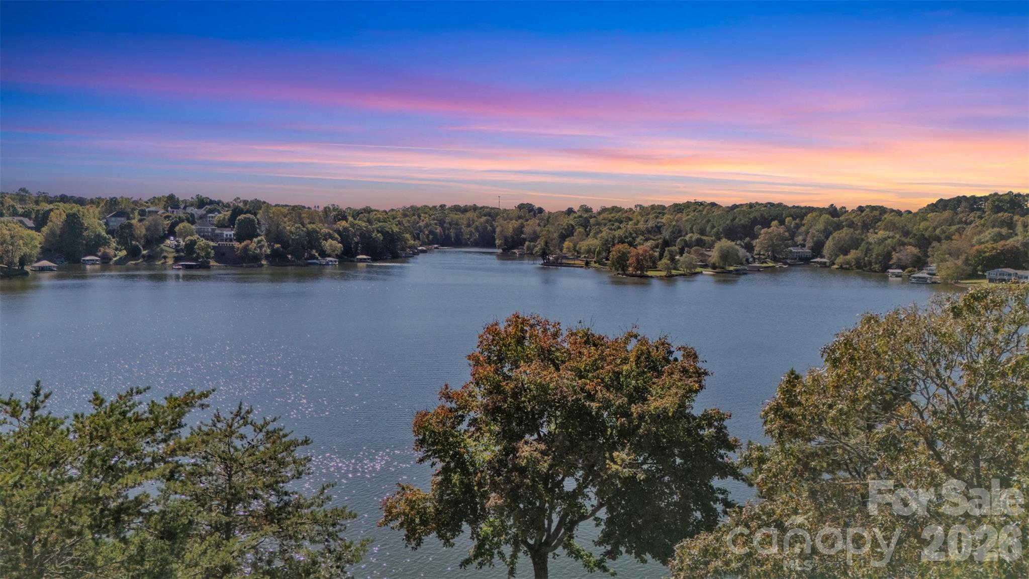 Panoramic lake view with wooded shores and homes beneath a colorful sunset sky.