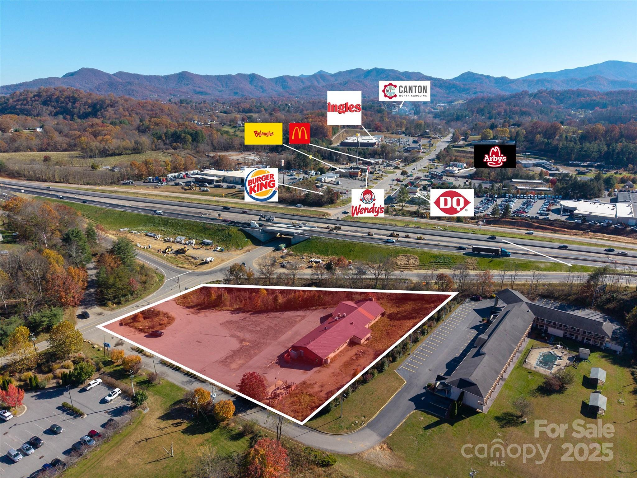 Aerial view of a commercial parcel outlined beside a highway, with a red-roofed building and mountains beyond.