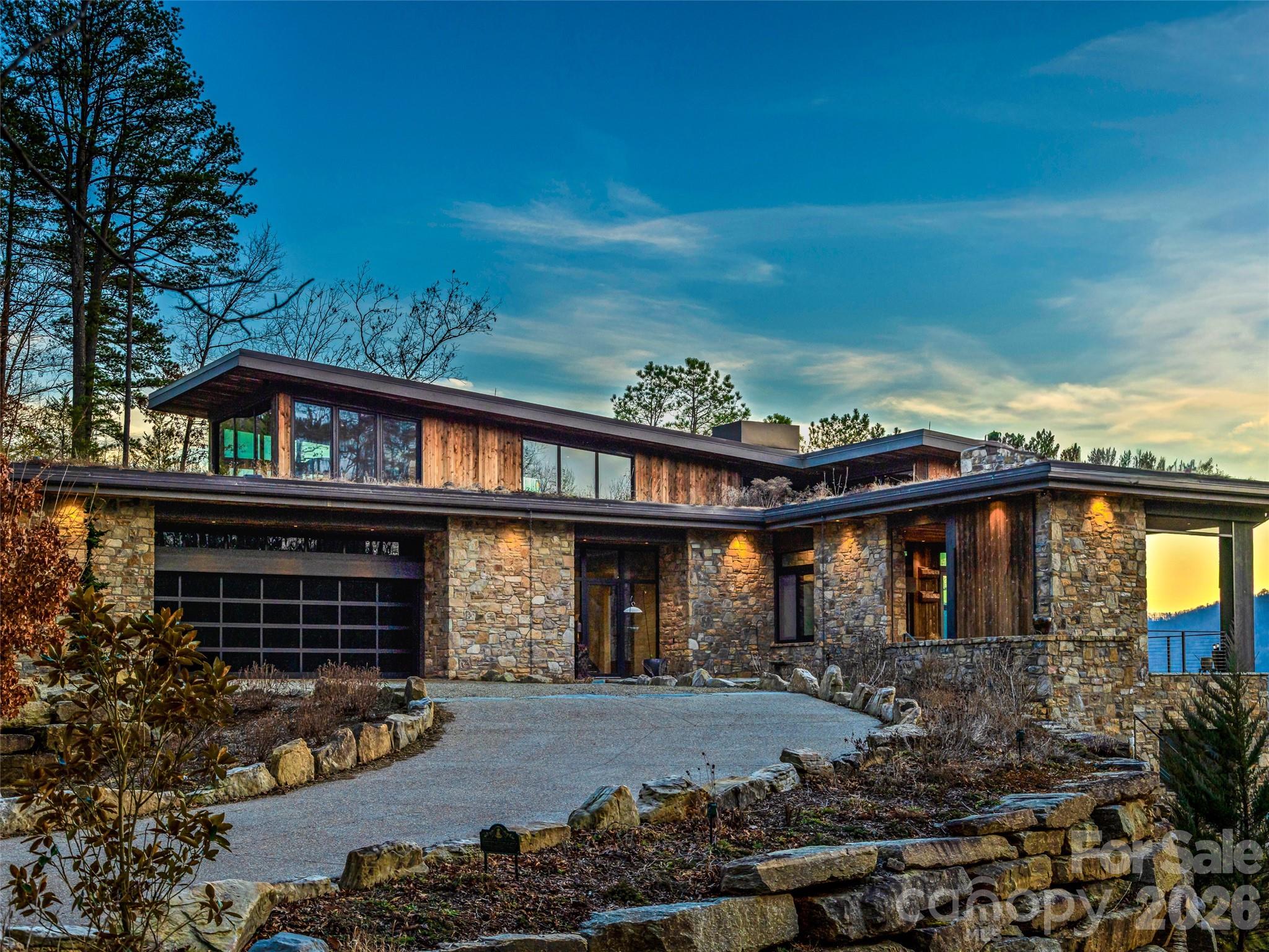 Modern stone-and-timber home with broad windows, a curved driveway, and water visible beside it.
