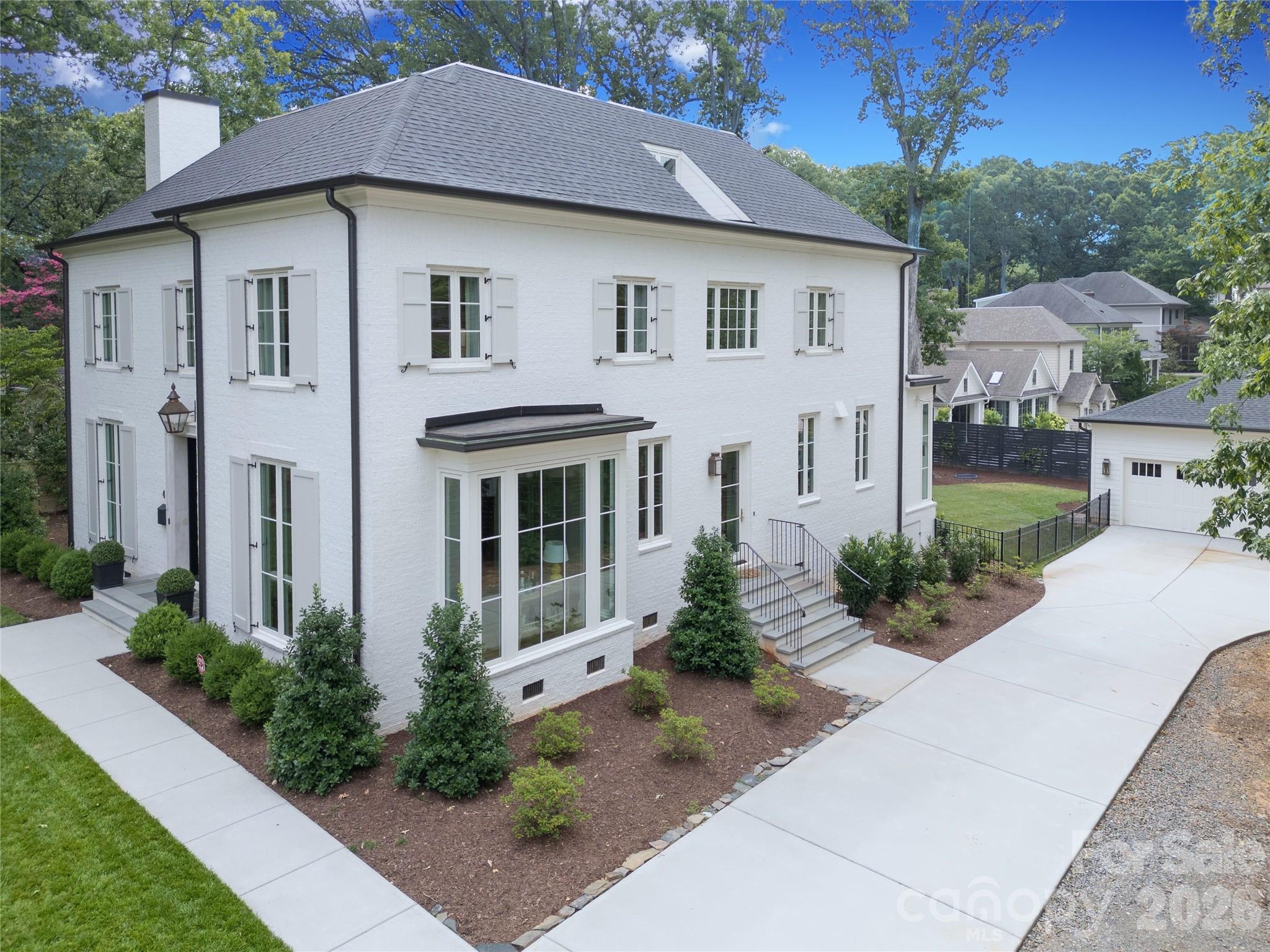 White brick home with gray roof, landscaped shrubs, and a broad driveway beside a detached garage.