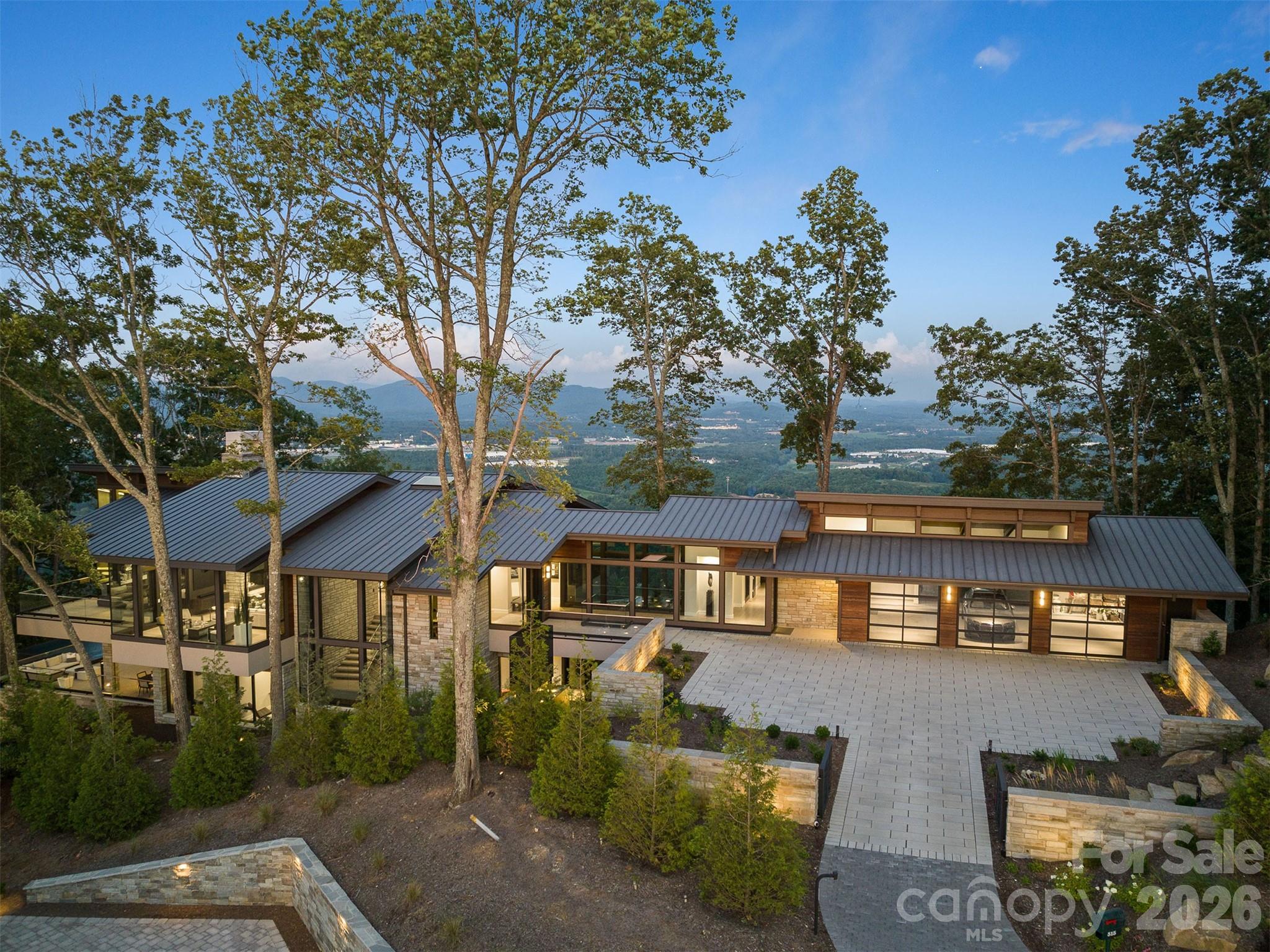 Modern stone-and-wood home with a broad metal roof, framed by trees and overlooking distant mountains.