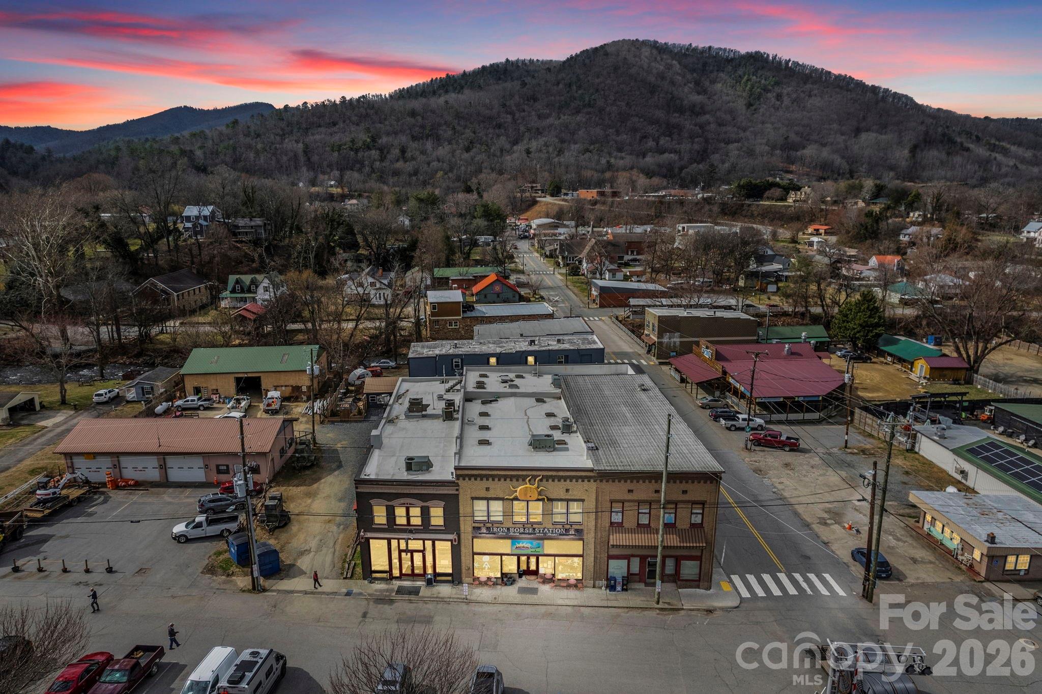 Aerial view of a small mountain town beneath a wooded ridge and pink dusk sky.