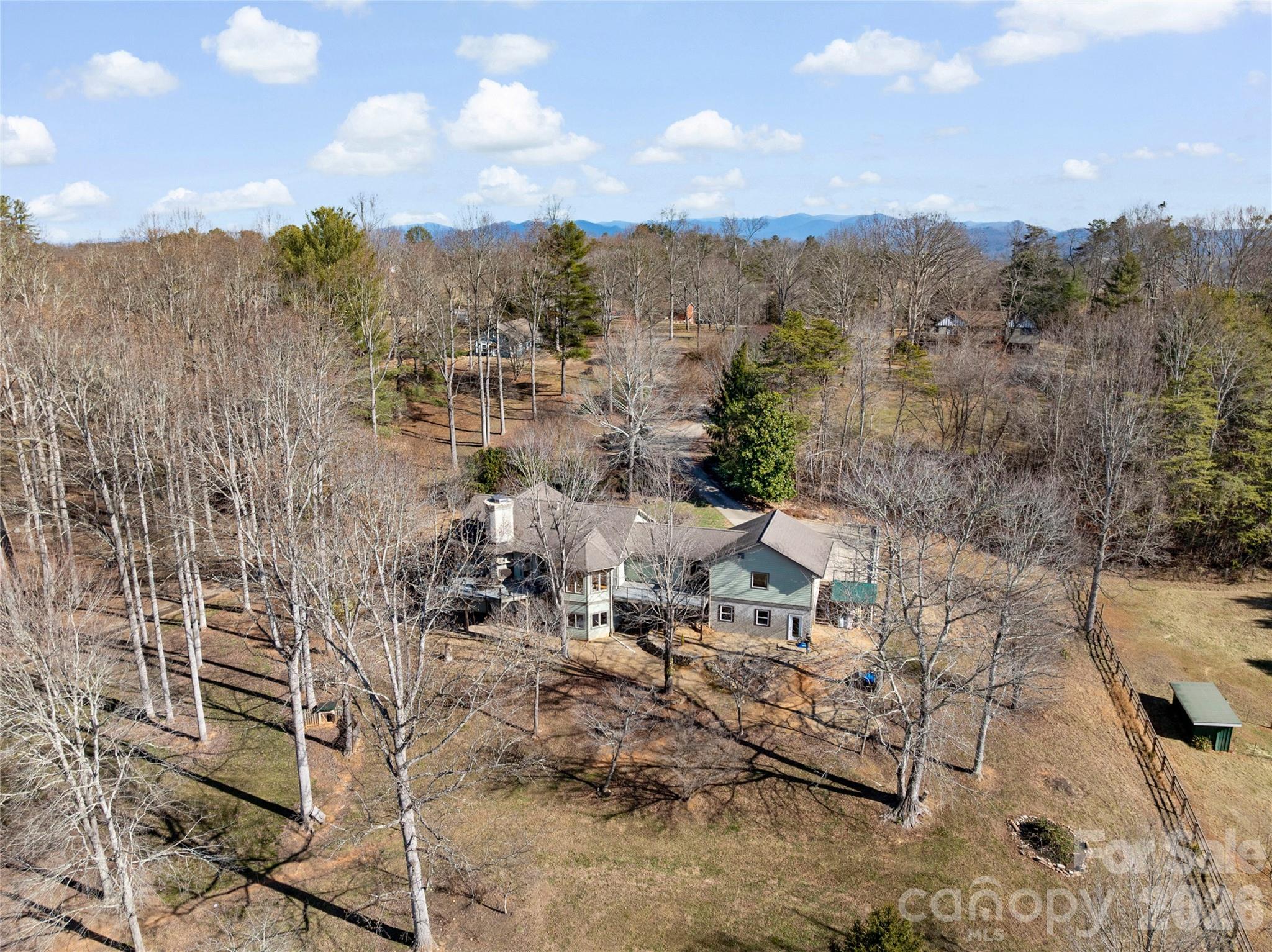 Aerial view of a home on a wooded lot, with a fenced lawn and distant mountain ridge.