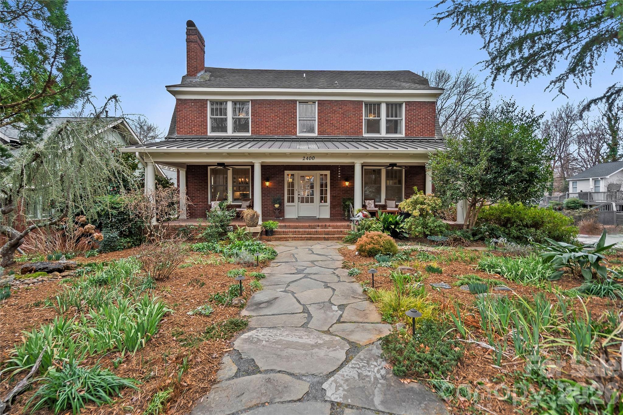 Two-story brick house with a covered porch and flagstone path through landscaped gardens.