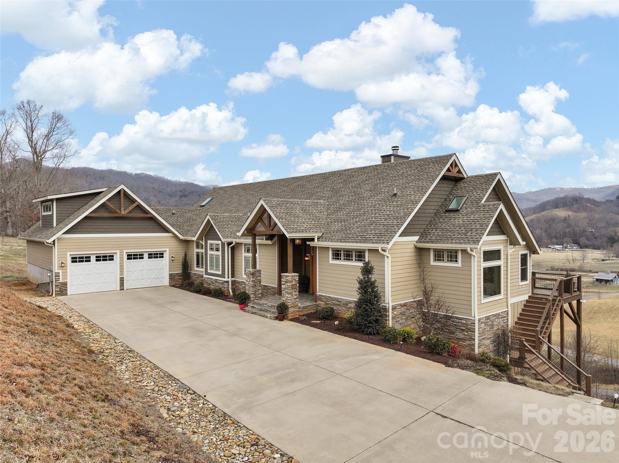Large beige home with multiple gables, two-car garage, and raised deck, set against rolling hills.