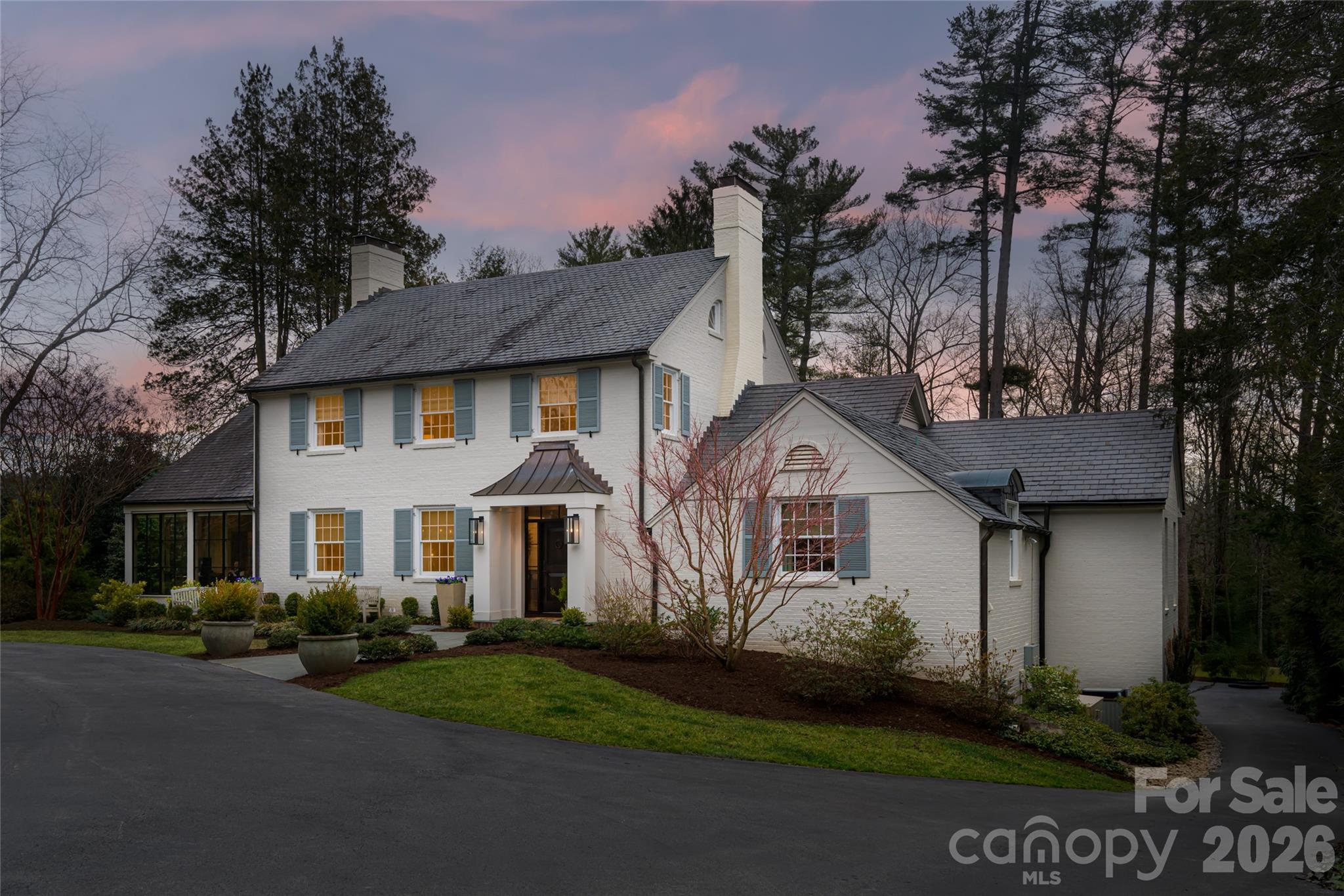 White brick two-story home with blue shutters and landscaped grounds, surrounded by trees at dusk.