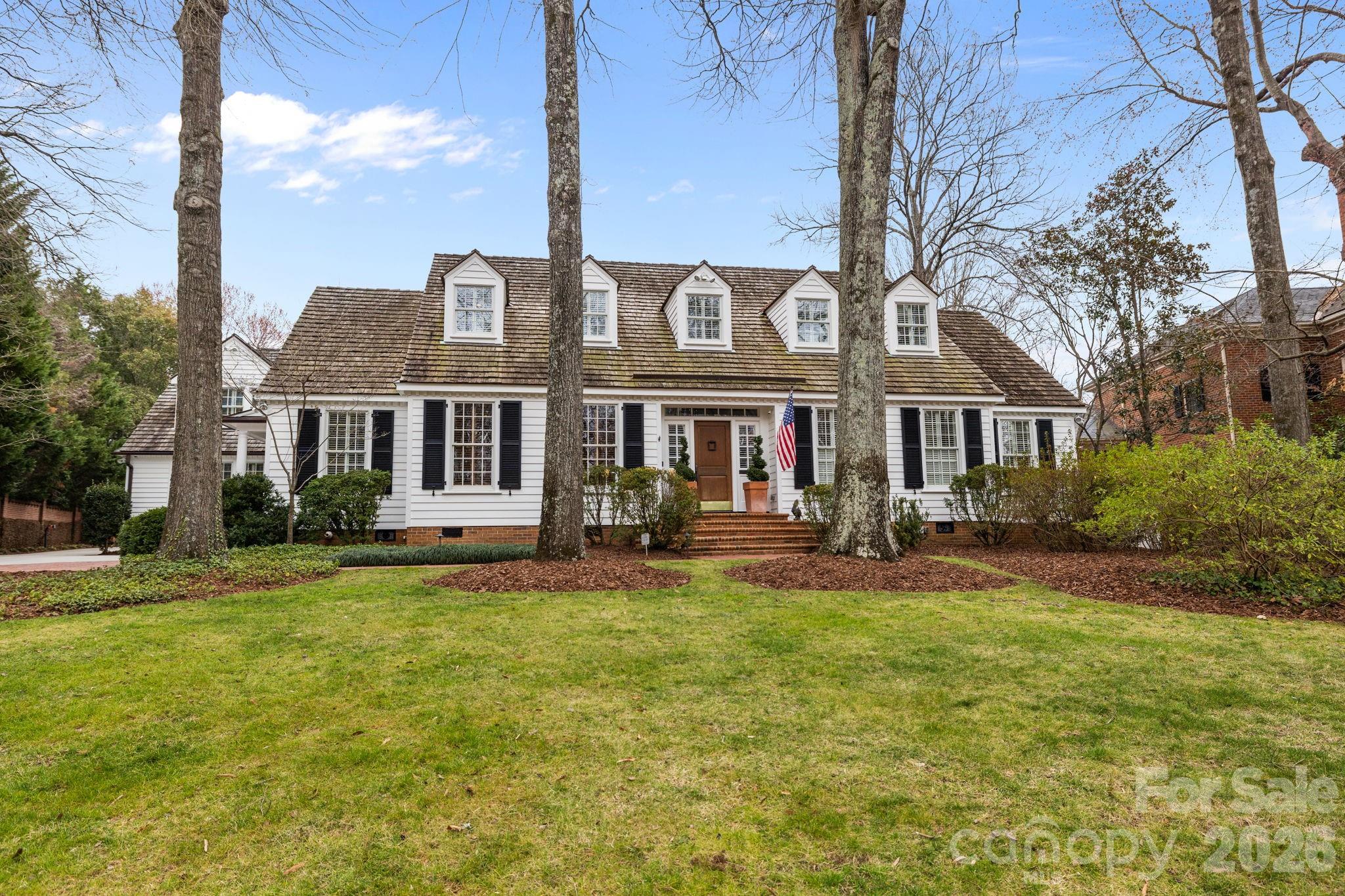 White clapboard house with five dormers, framed by tall trees and a broad front lawn.