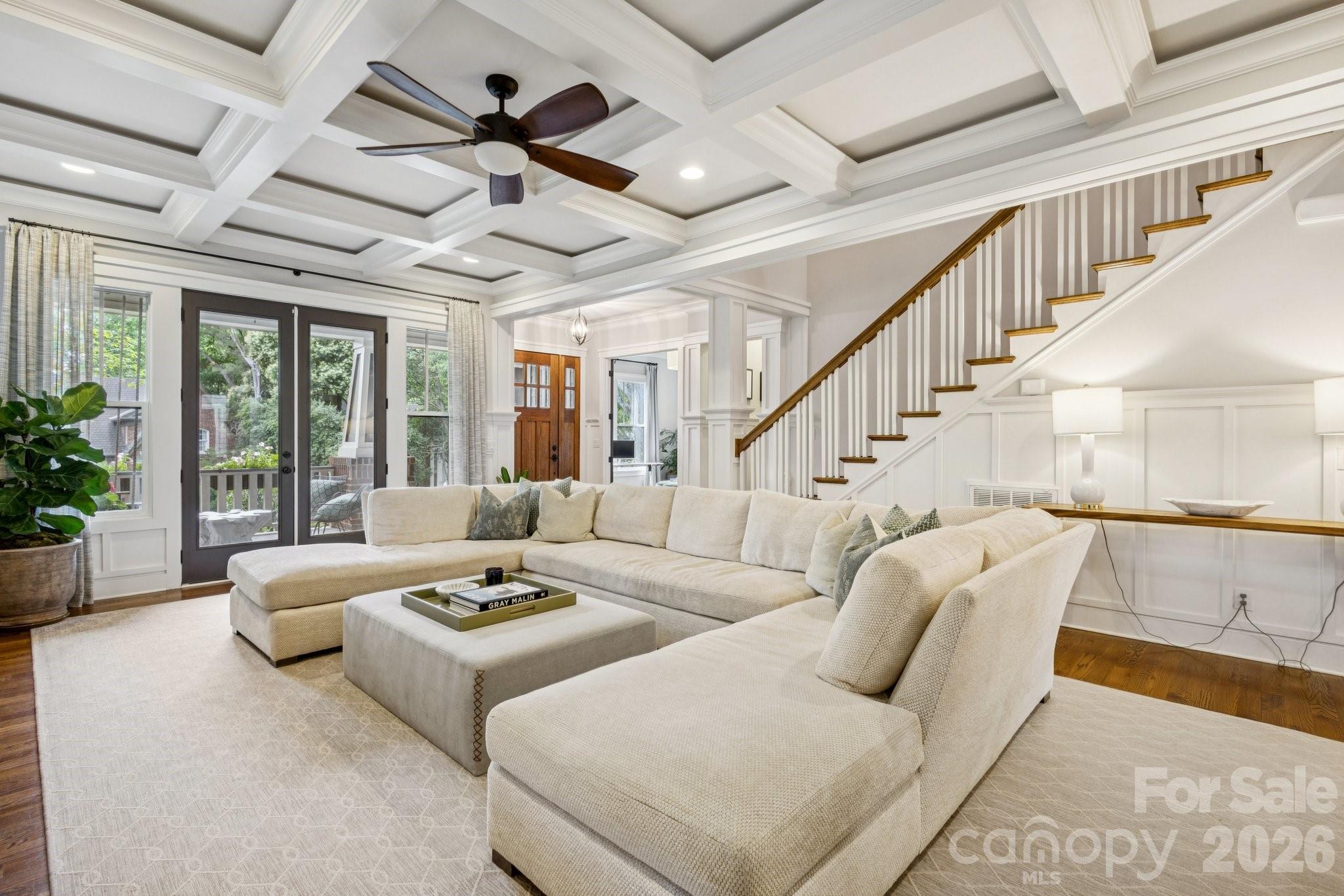 Furnished living room with a cream sectional, coffered ceiling, and staircase with a wood handrail.