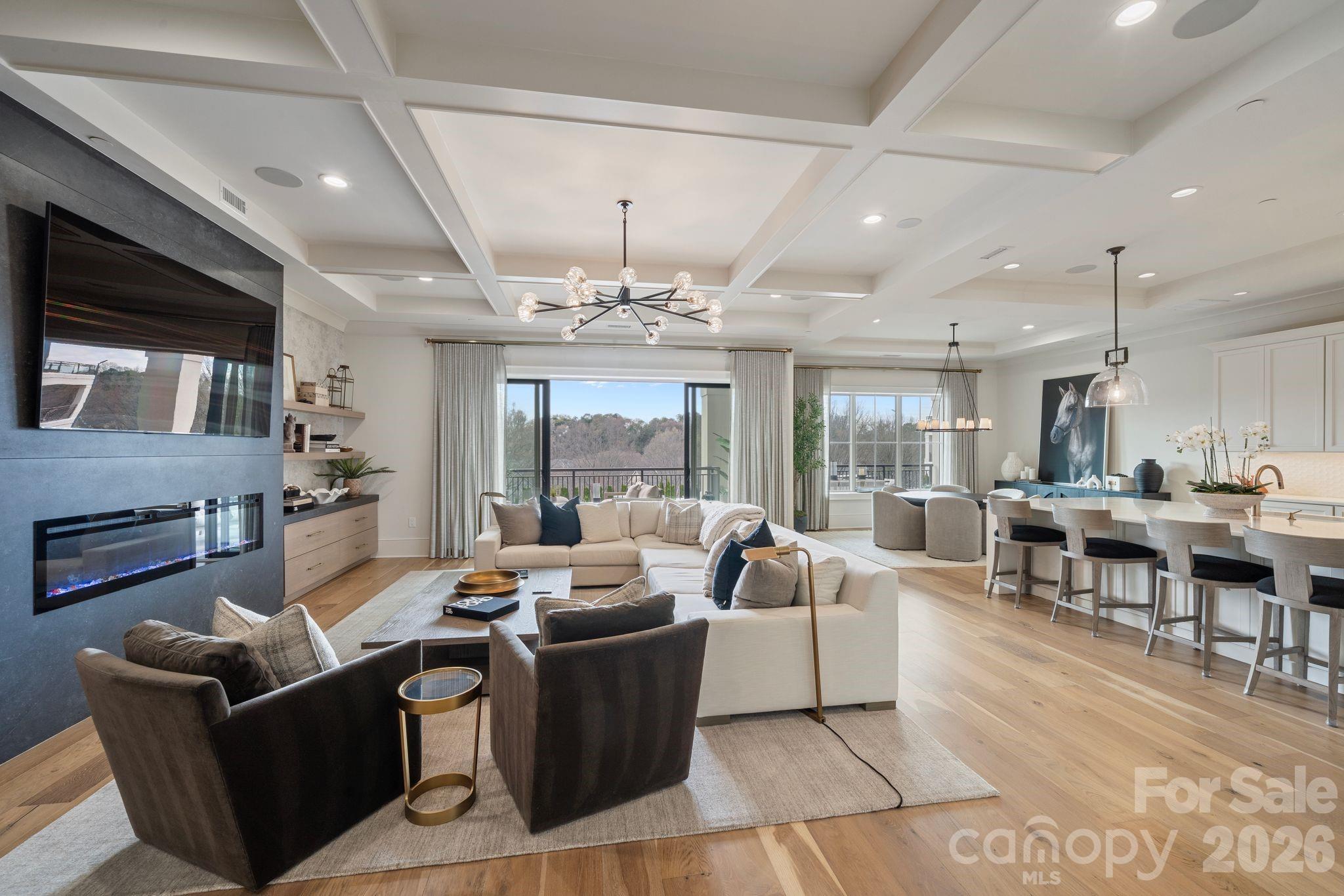 Open-plan living and dining room with a coffered ceiling, fireplace, and wooded balcony view.