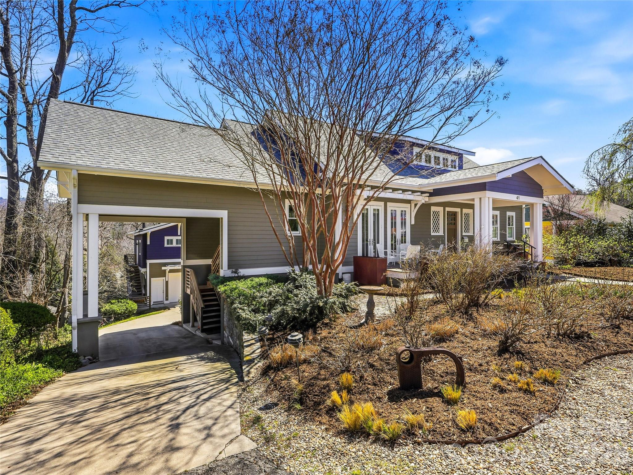 Gray-sided home with a covered porch, open carport, and gravel garden beneath a leafless tree.