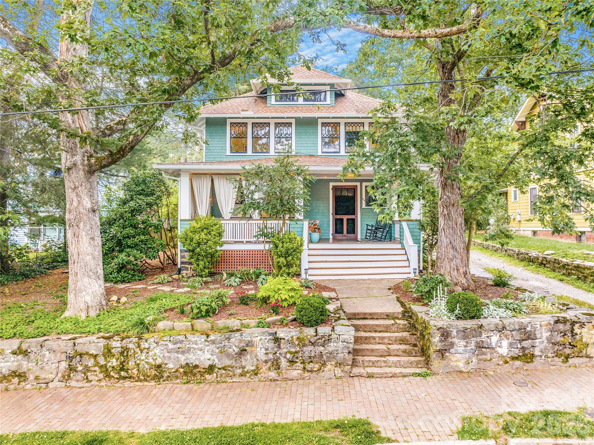 Turquoise two-story home with a broad front porch, stone retaining wall, and mature trees.