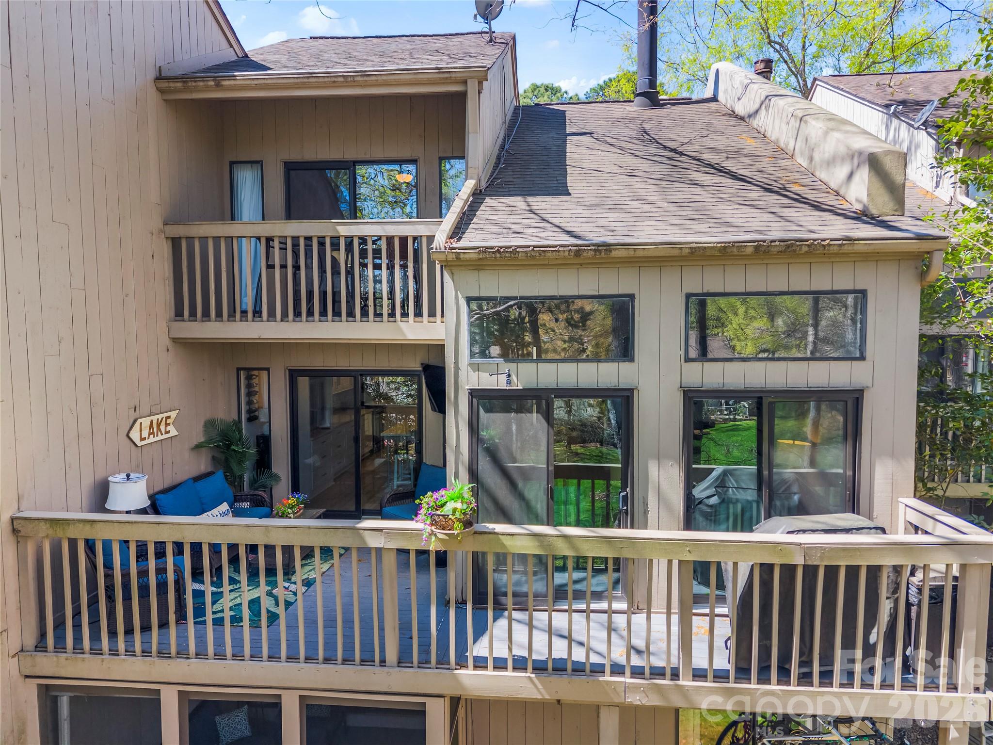 Multi-level beige home with stacked decks, blue patio seating, and a sign reading “LAKE.”