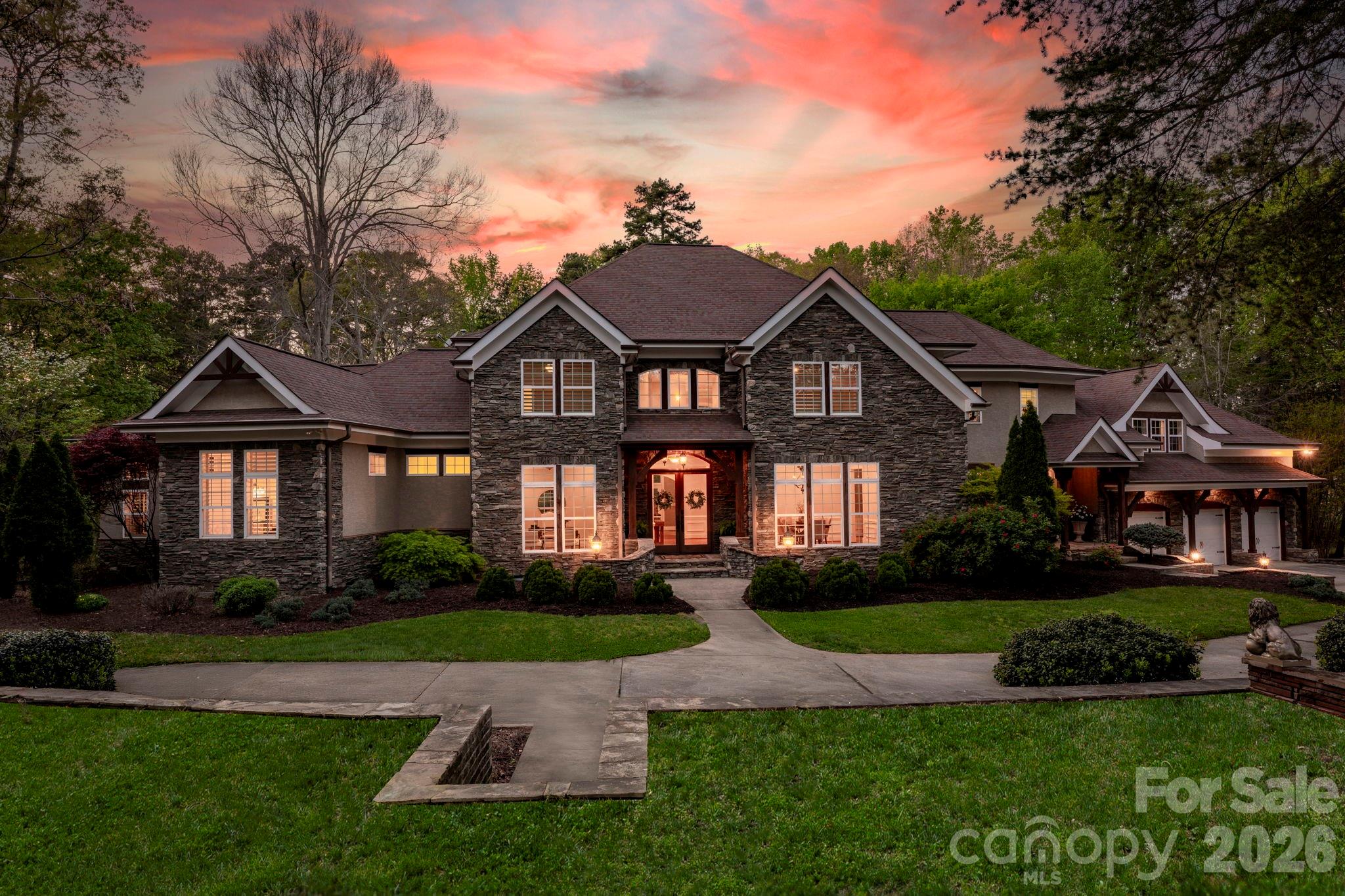 Two-story stone house with warmly lit windows, landscaped lawn, and a colorful sunset sky.