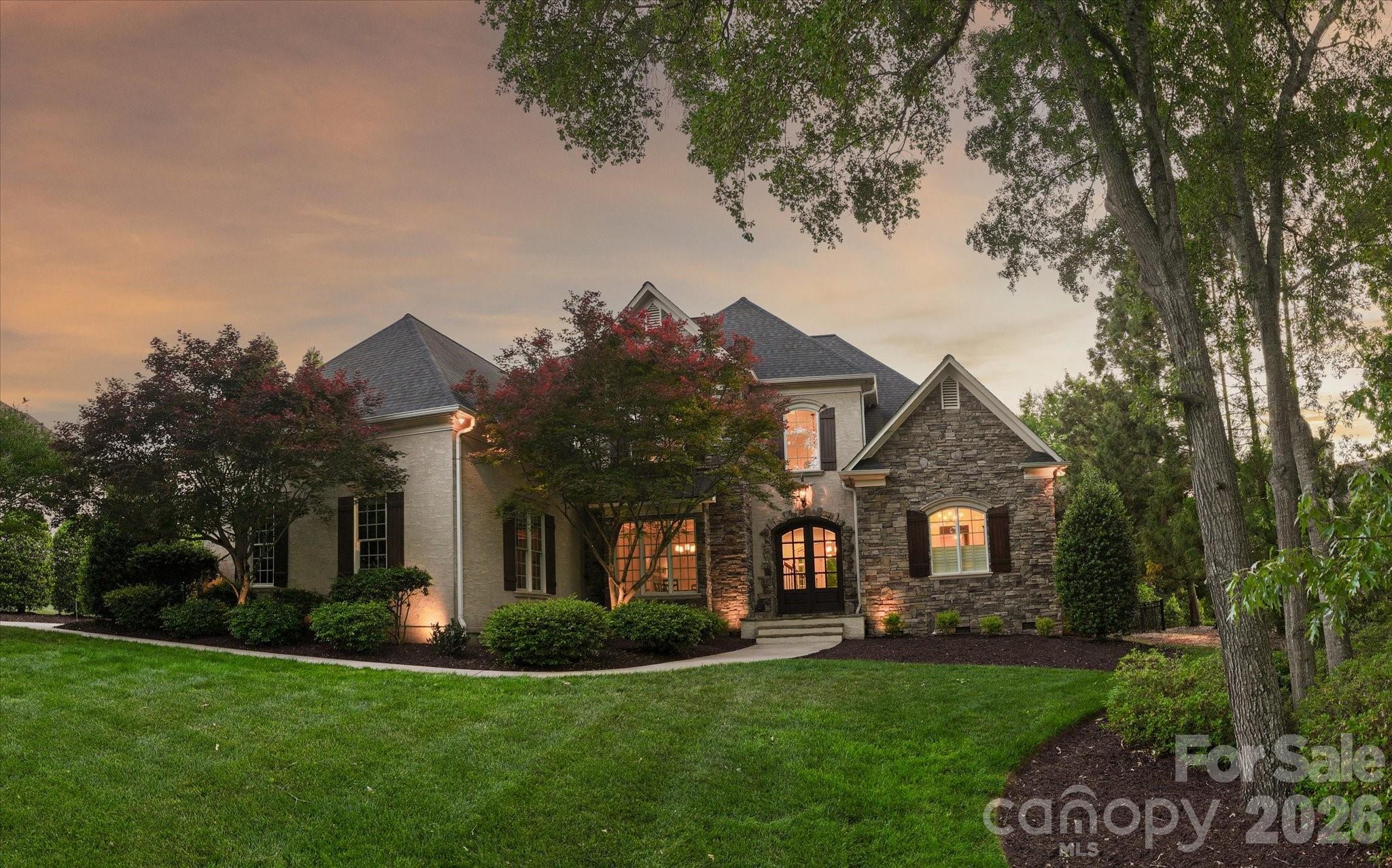 Two-story stone-and-siding home with illuminated windows, mature trees, and a broad lawn at dusk.