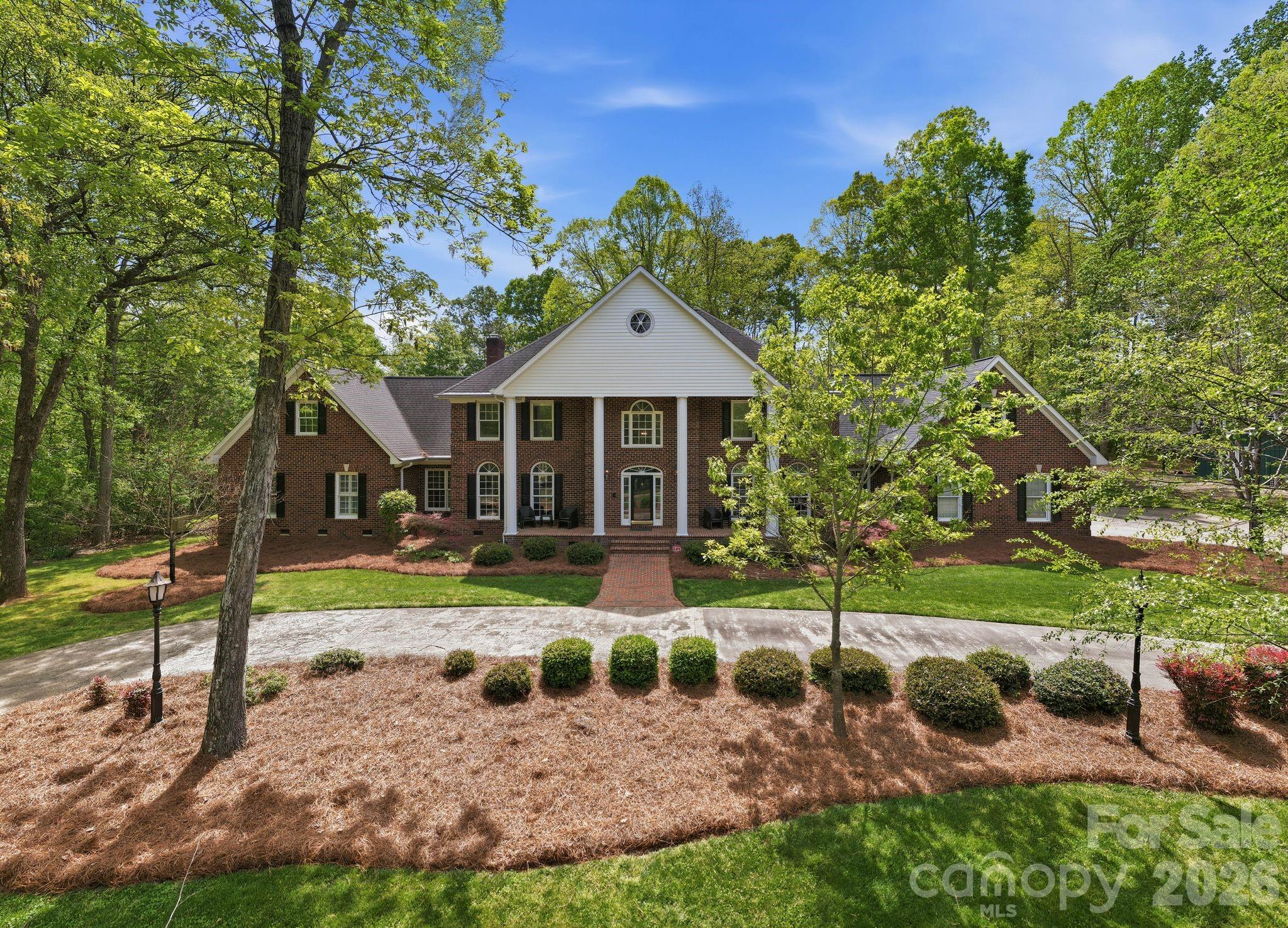 Two-story brick home with a prominent white gable, mature trees, and a landscaped front lawn.