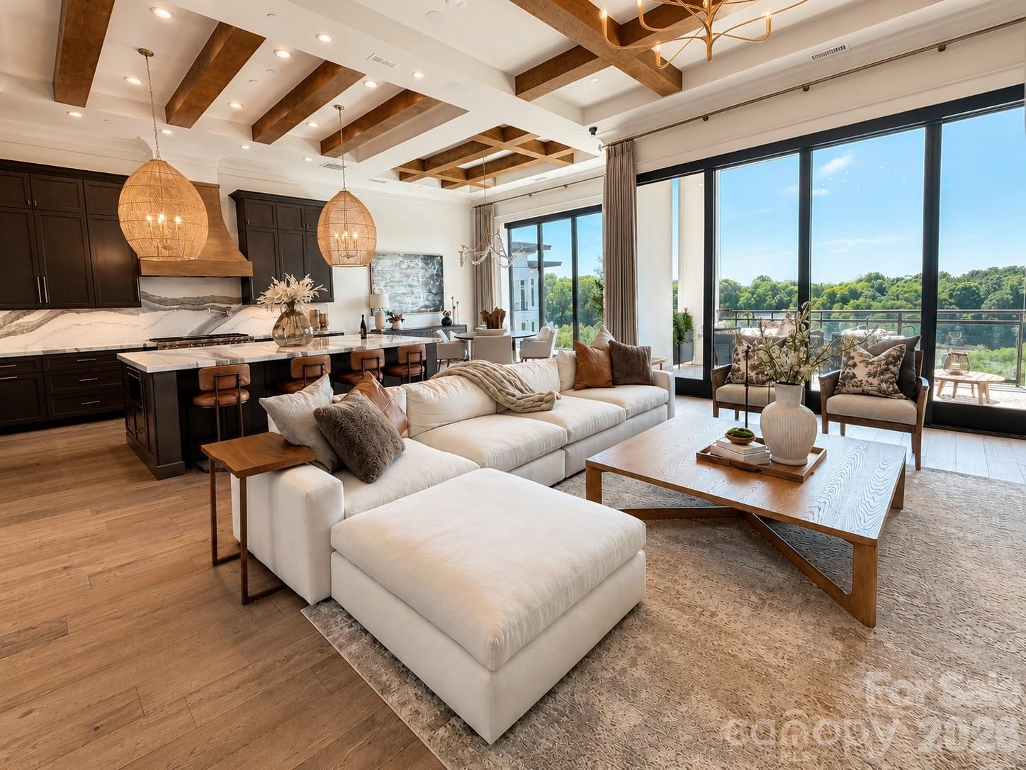 Open-plan condo living room with a white sectional, marble kitchen island, wood ceiling beams, and leafy views.