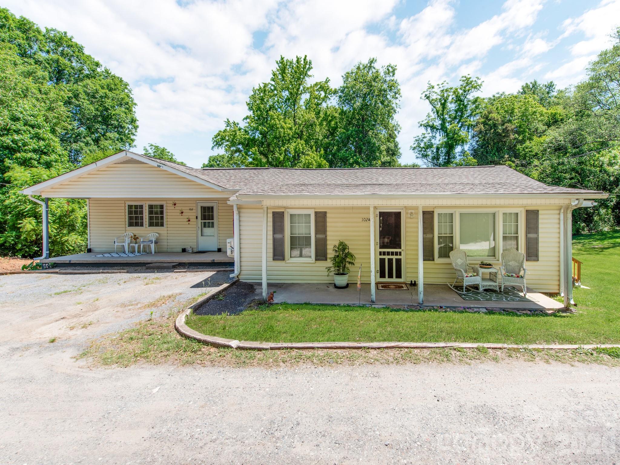 Pale yellow single-story house with two covered porches, rocking chairs, and a gravel driveway.