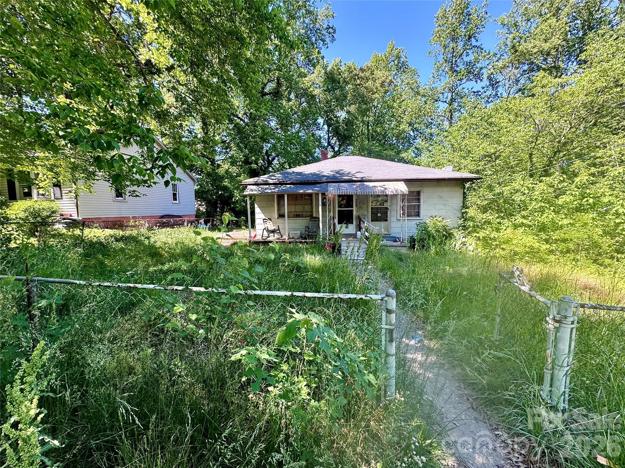 Single-story house with a covered porch behind an overgrown, fenced yard and mature trees.