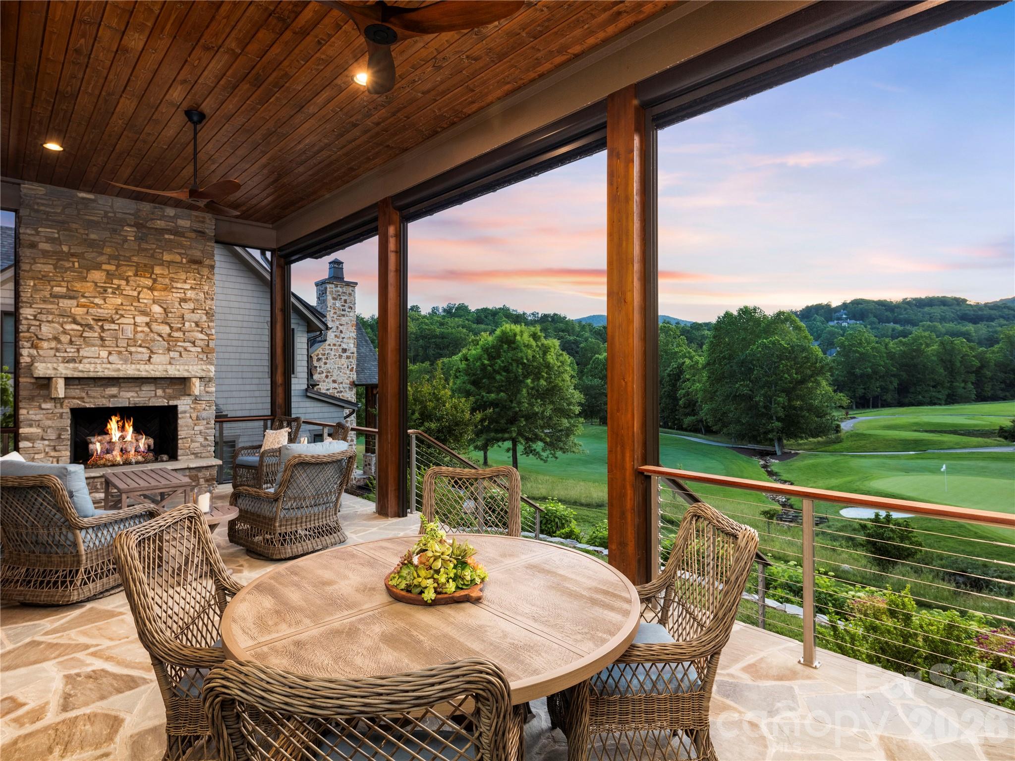 Covered patio with a stone fireplace and wicker seating overlooking a golf course at sunset.