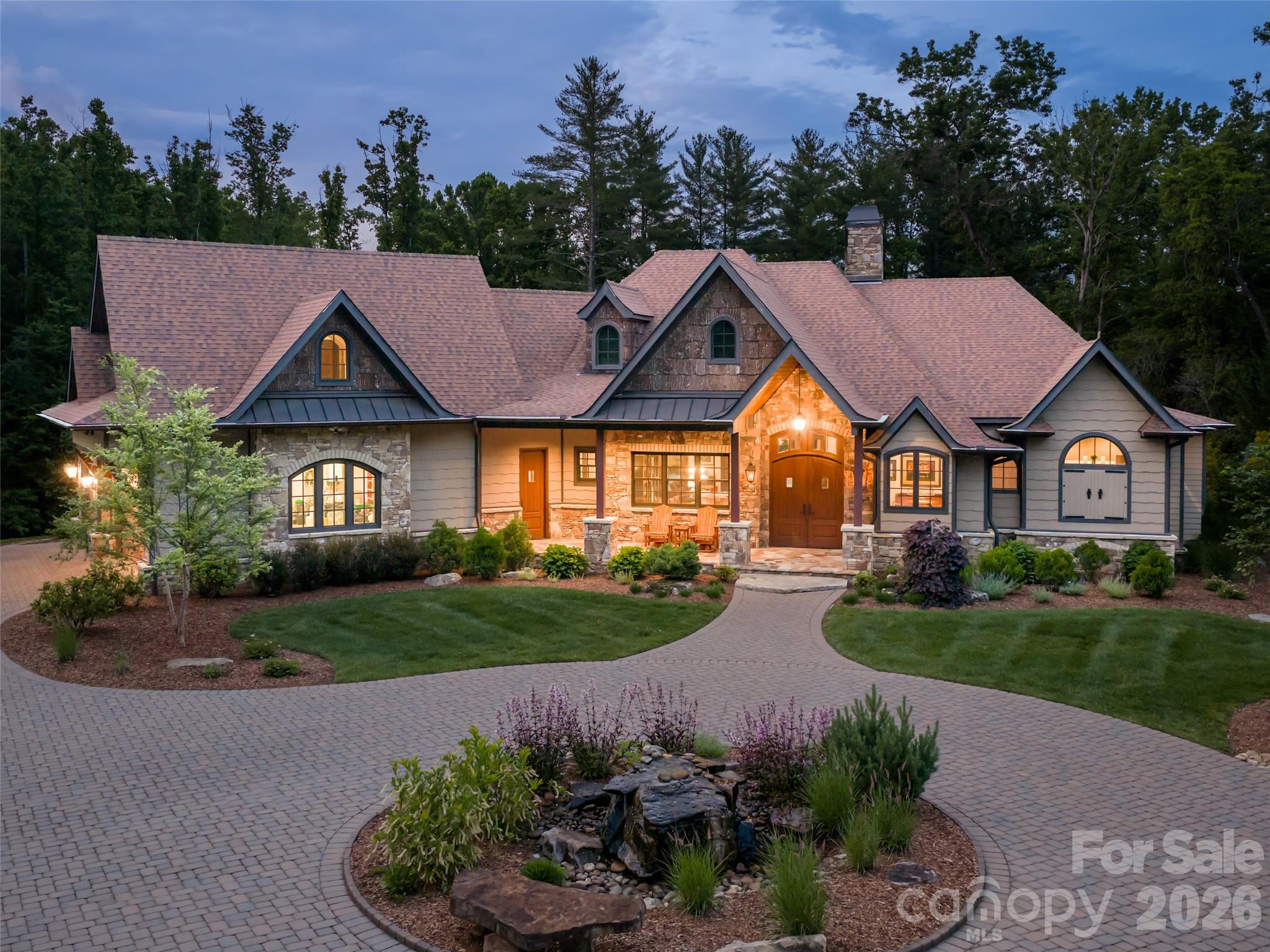 Craftsman-style home with a stone entrance, landscaped lawn, curved paver driveway, and central rock garden at dusk.