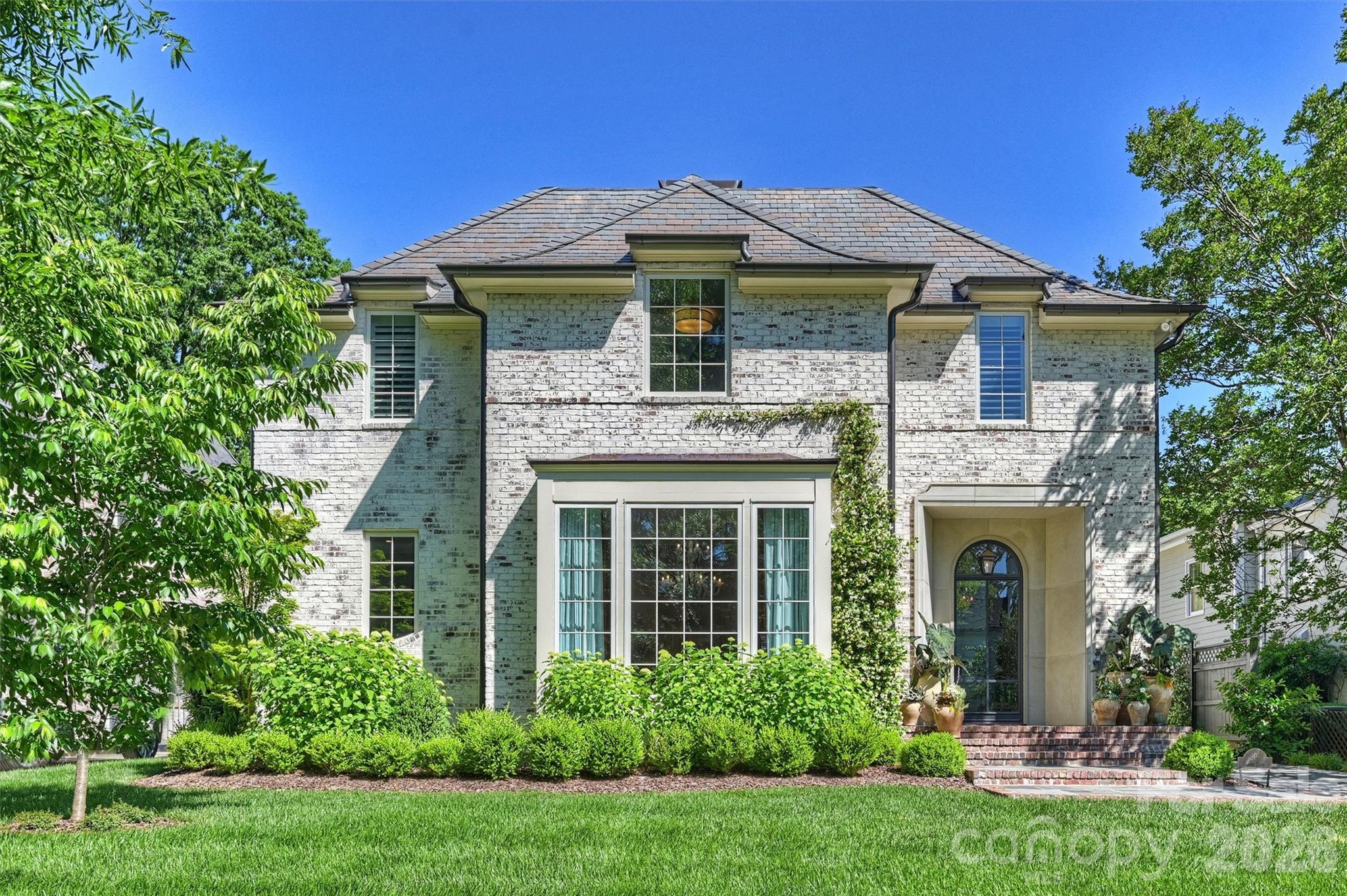 Two-story whitewashed brick home with an arched entrance, leafy landscaping, and a broad front lawn.