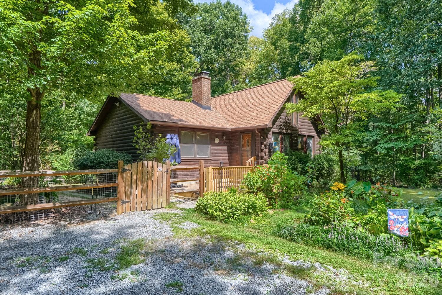 Log cabin surrounded by trees and gardens, with a wooden fence and gravel driveway.