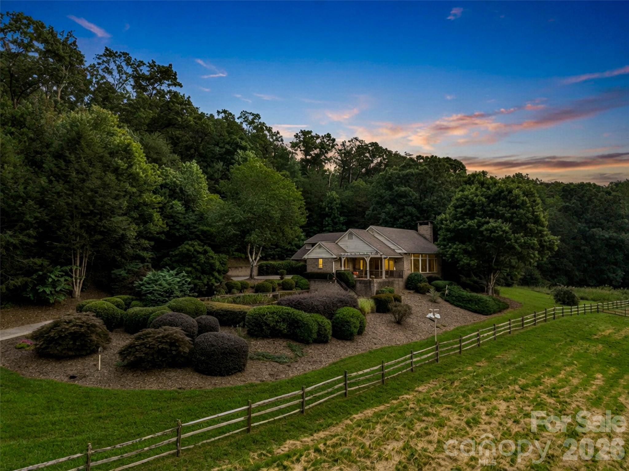 Wooded home with landscaped grounds, a split-rail fence, and glowing windows at dusk.