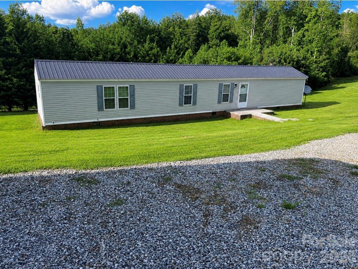 Manufactured home with a dark metal roof, grassy lawn, gravel driveway, and wooded backdrop.